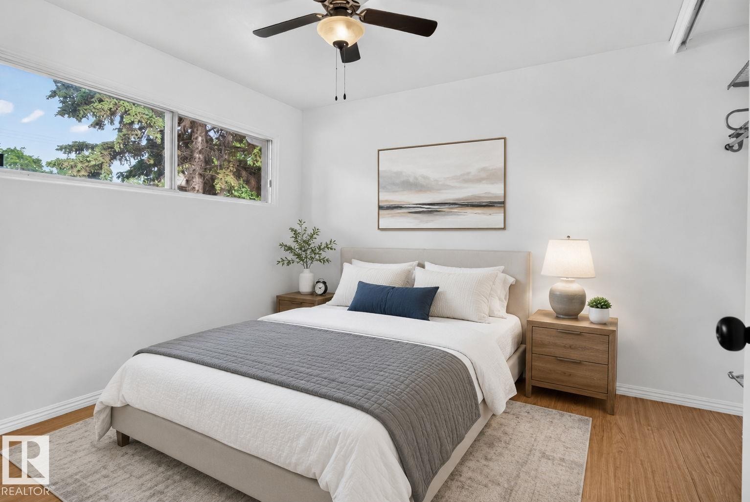 Bedroom featuring wide horizontal windows, a ceiling fan with integrated lighting, wood-finish flooring, a light-colored headboard, and white baseboards - 12952 114 Street Nw, Edmonton, AB - Indoor Photo Showing Bedroom