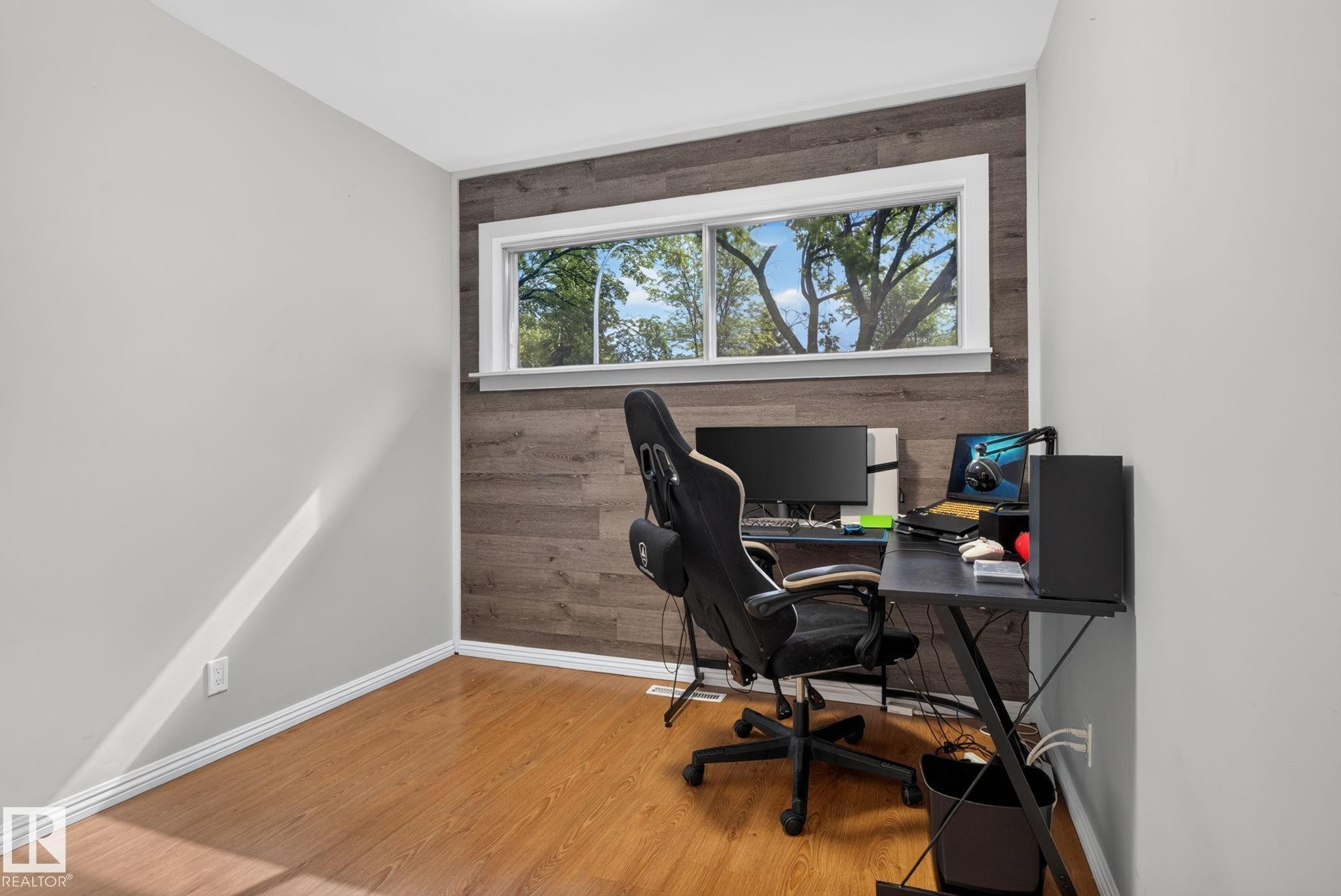 Room featuring a wide window, wood-finish accent wall, light-toned wall paint, white trim, and wood-finish flooring - 12952 114 Street Nw, Edmonton, AB