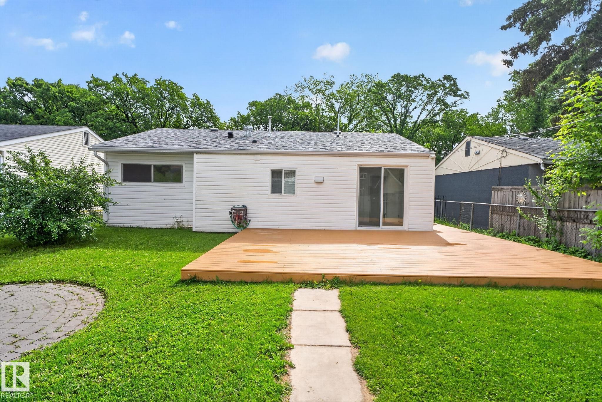 Expansive backyard featuring a large wood-finish deck, sliding glass door access, a circular stone patio, green lawn, and mature trees - 12952 114 Street Nw, Edmonton, AB