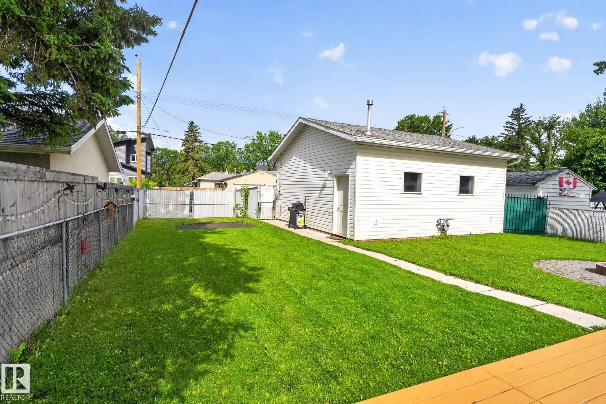 Expansive fenced backyard featuring a detached garage with vinyl siding, a concrete pathway, a circular paver patio, and mature perimeter trees - 12952 114 Street Nw, Edmonton, AB