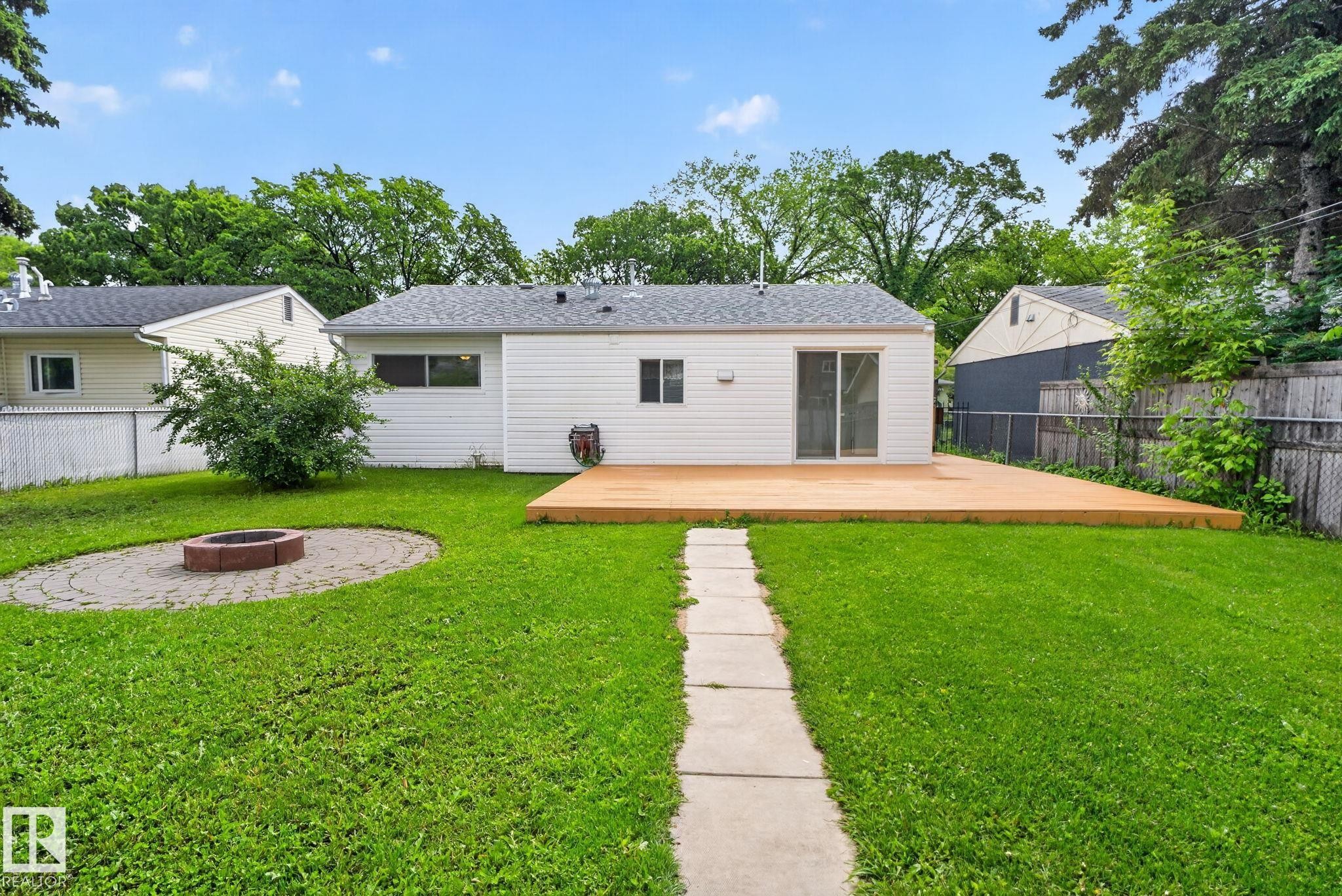 Expansive backyard featuring a central lawn, a paver patio with a fire pit, a concrete stepping stone path, a spacious wood deck, and white siding - 12952 114 Street Nw, Edmonton, AB