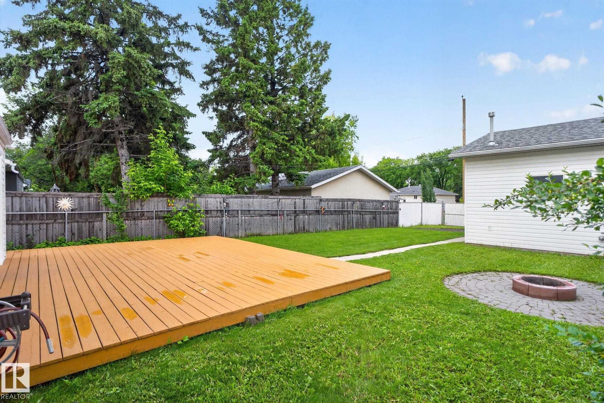 Spacious backyard featuring a large composite deck, a circular paver patio with a fire pit, mature perimeter trees, a wooden fence, and an exterior building with white siding - 12952 114 Street Nw, Edmonton, AB