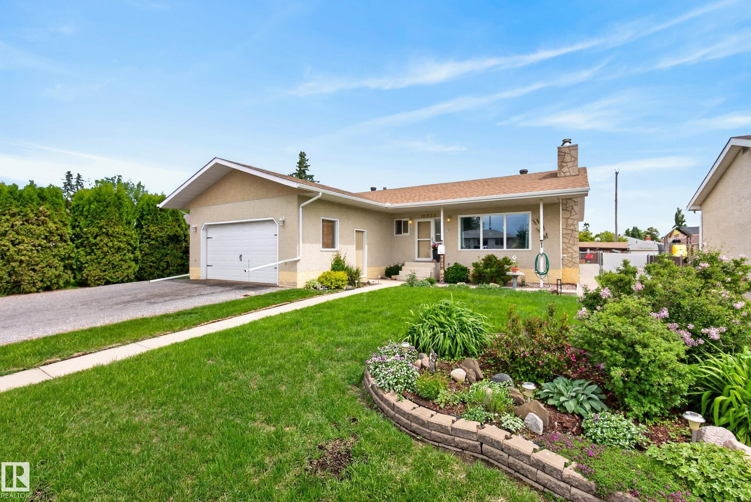 Exterior featuring a stucco finish, attached garage, paved driveway, landscaped front yard with a stone-edged garden bed, and mature perimeter trees - 10935 152 Street, Edmonton, AB