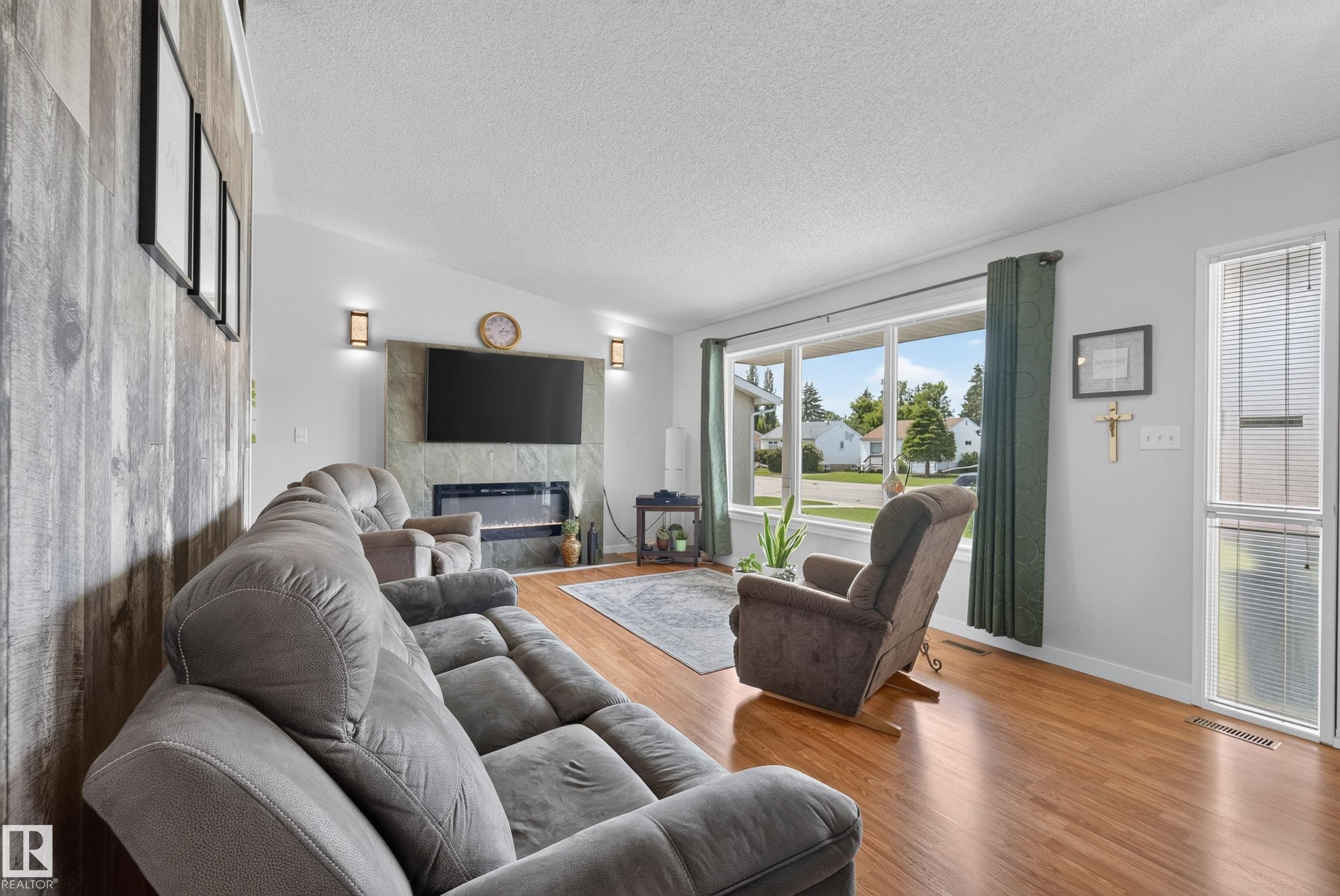 Living area featuring wood-finish flooring, a contemporary fireplace with stone surround, a prominent picture window, and a vaulted ceiling with textured finish - 10935 152 Street, Edmonton, AB