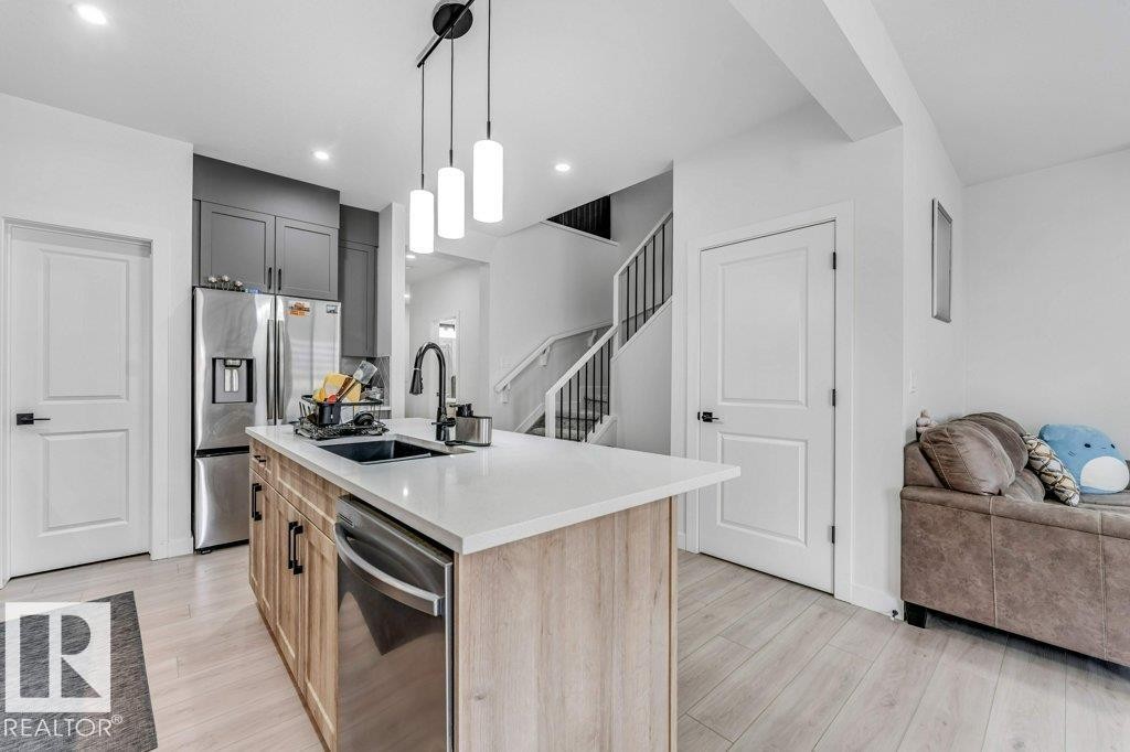 Modern kitchen featuring a light wood-finish island with a white countertop and undermount sink, stainless steel appliances, dark gray upper cabinetry, and wood-finish flooring - 423 28 Street, Edmonton, AB