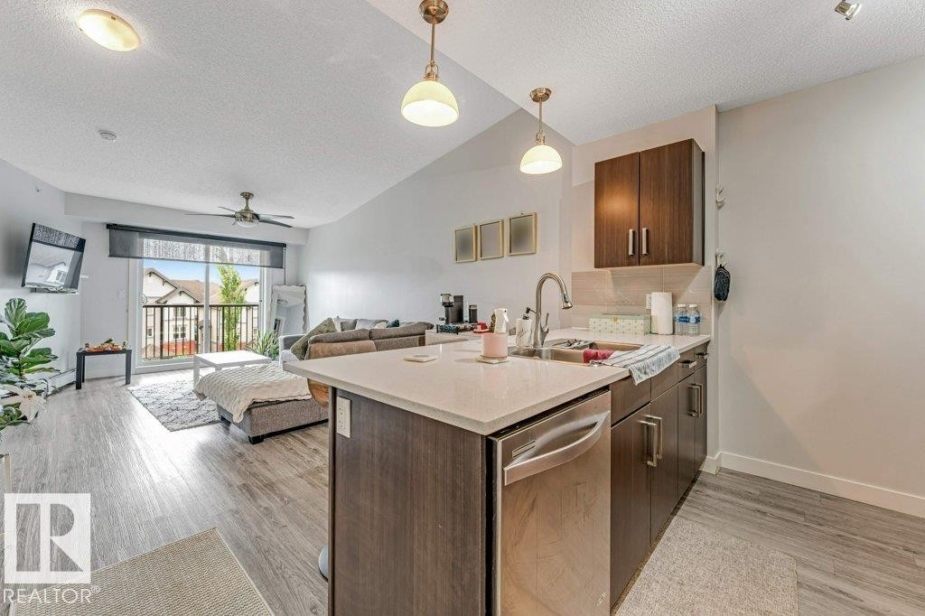 Open-concept living space featuring a modern kitchen island with an integrated stainless steel dishwasher, wood-finish flooring, a sliding glass door to an exterior balcony, and a ceiling fan - 401 667 Watt Boulevard, Edmonton, AB