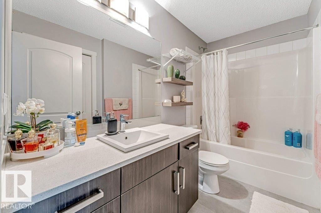 Modern bathroom featuring a wood-finish vanity with a light-toned countertop, an integrated rectangular sink, a bathtub with a shower, and light gray floor tiles - 401 667 Watt Boulevard, Edmonton, AB