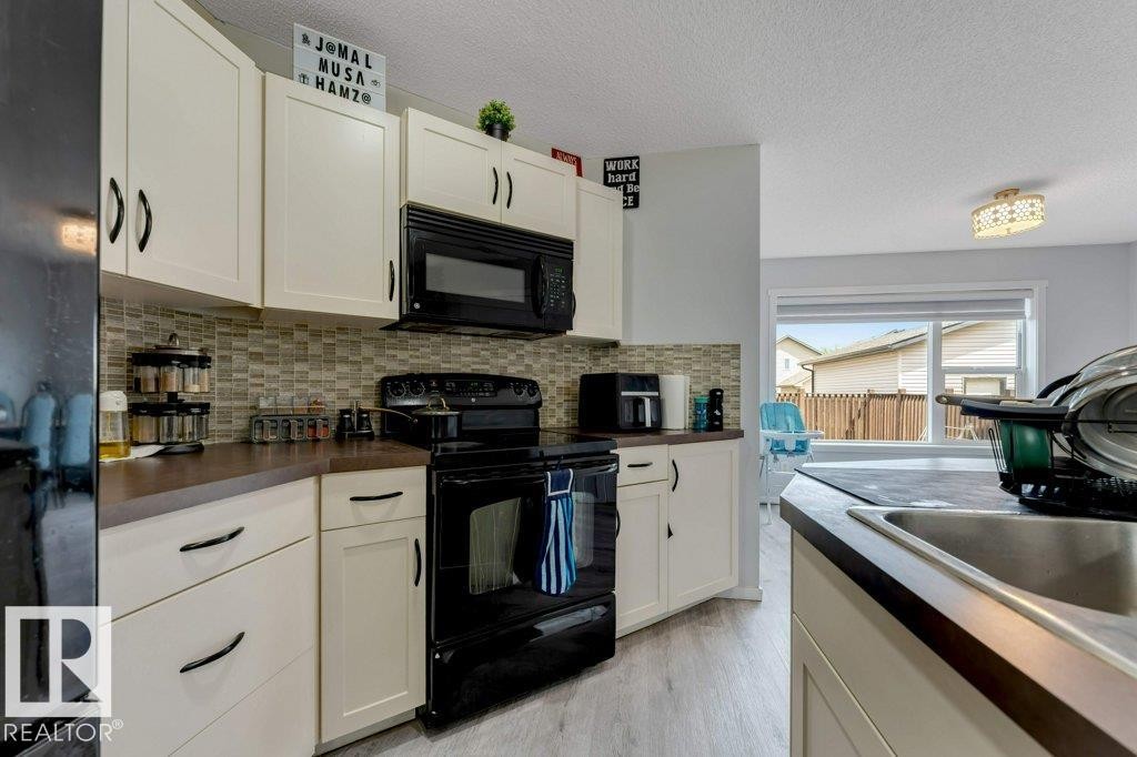 Kitchen featuring cream-toned cabinetry with dark hardware, dark countertops, a mosaic tile backsplash, black appliances, and light wood-finish flooring - 3511 15A Street, Edmonton, AB