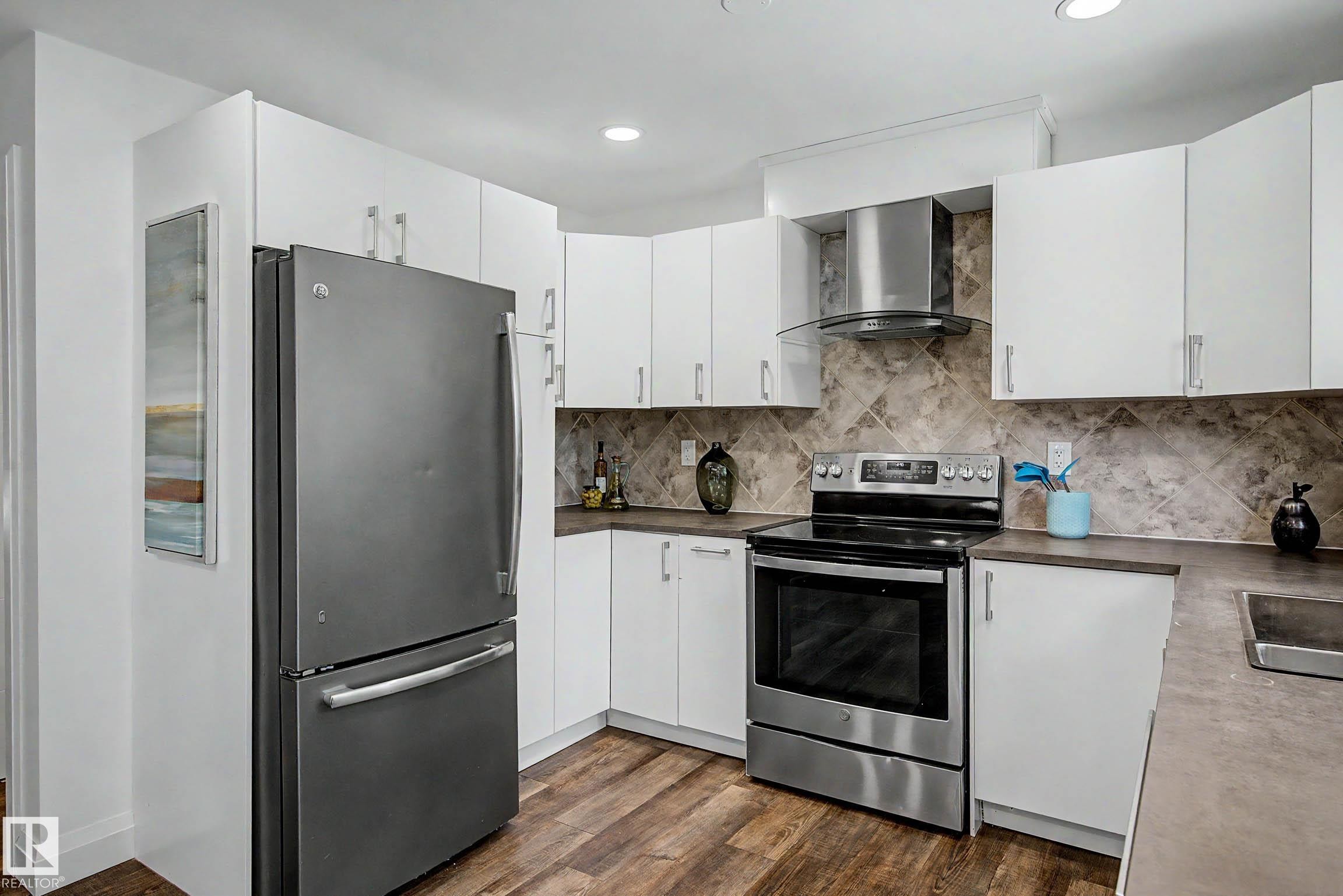 Kitchen featuring white cabinetry with silver hardware, wood-finish flooring, and a patterned tile backsplash - 3718 103B Street Nw, Edmonton, AB - Indoor Photo Showing Kitchen With Double Sink