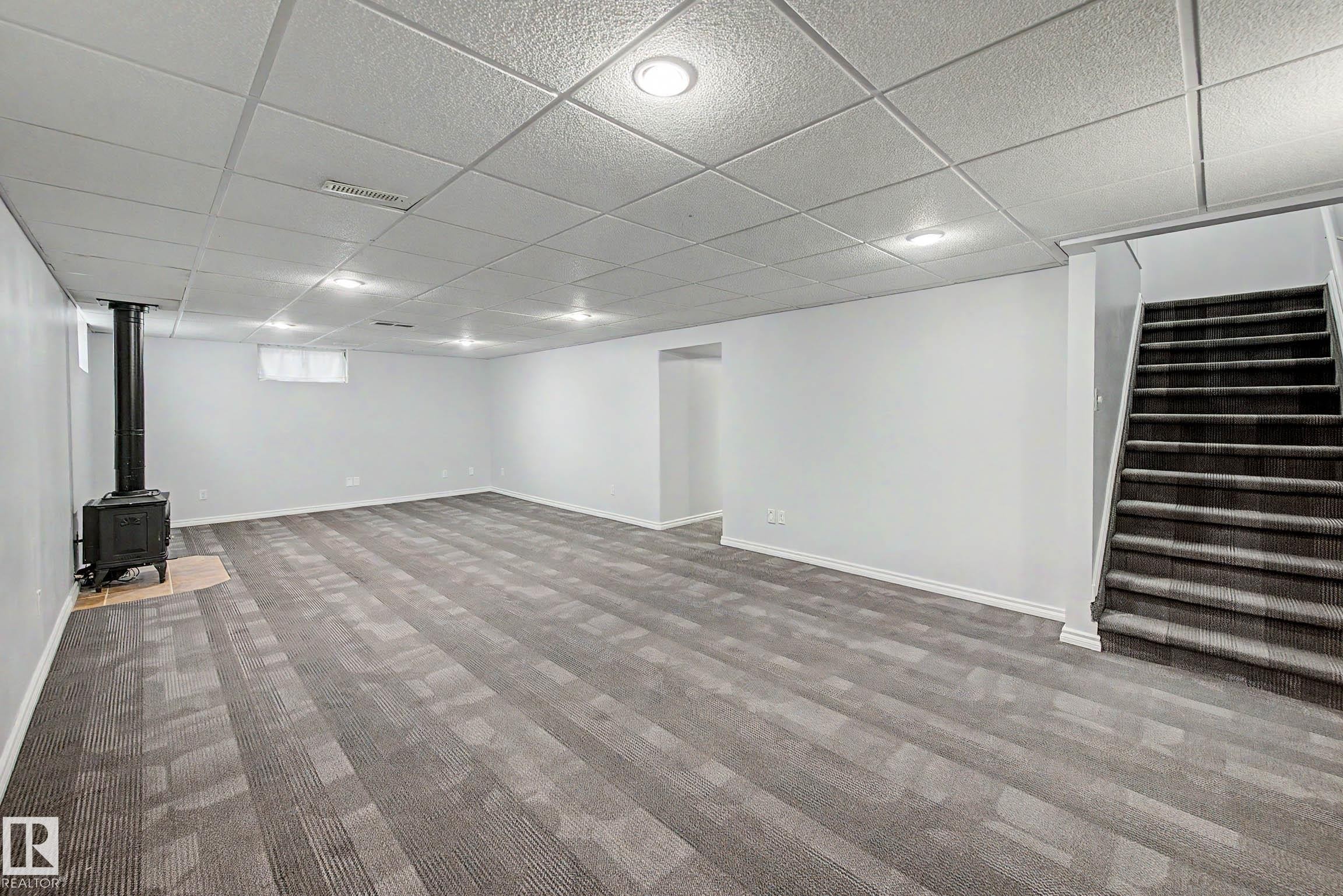 Spacious lower-level room featuring patterned carpet flooring, a black wood-burning stove on a tiled hearth, recessed lighting, and a drop ceiling - 3718 103B Street Nw, Edmonton, AB - Indoor Photo Showing Other Room