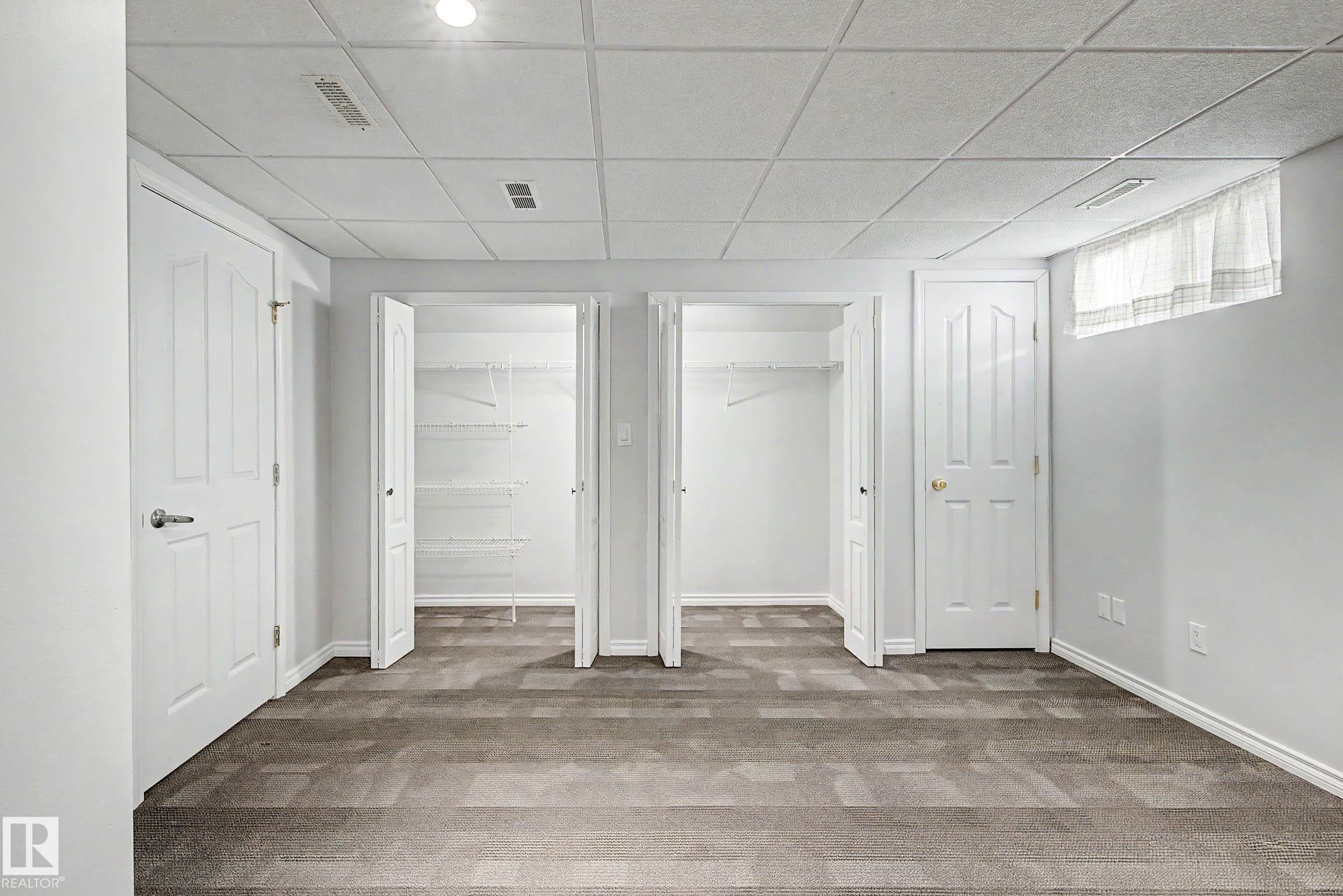 Carpeted room featuring two white paneled doors, two open closets with wire shelving, a window with curtain, and a dropped ceiling with recessed lighting - 3718 103B Street Nw, Edmonton, AB - Indoor Photo Showing Other Room