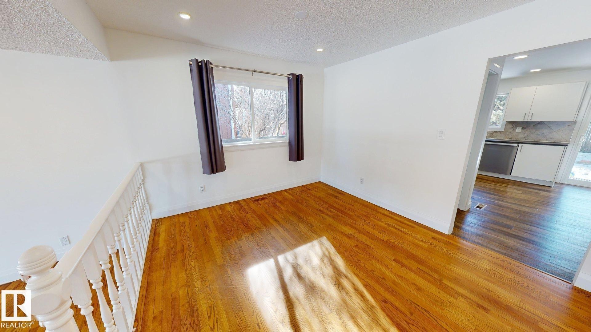 Hardwood flooring throughout a bright room featuring a window with dark curtains and recessed ceiling lights - 3718 103B Street, Edmonton, AB