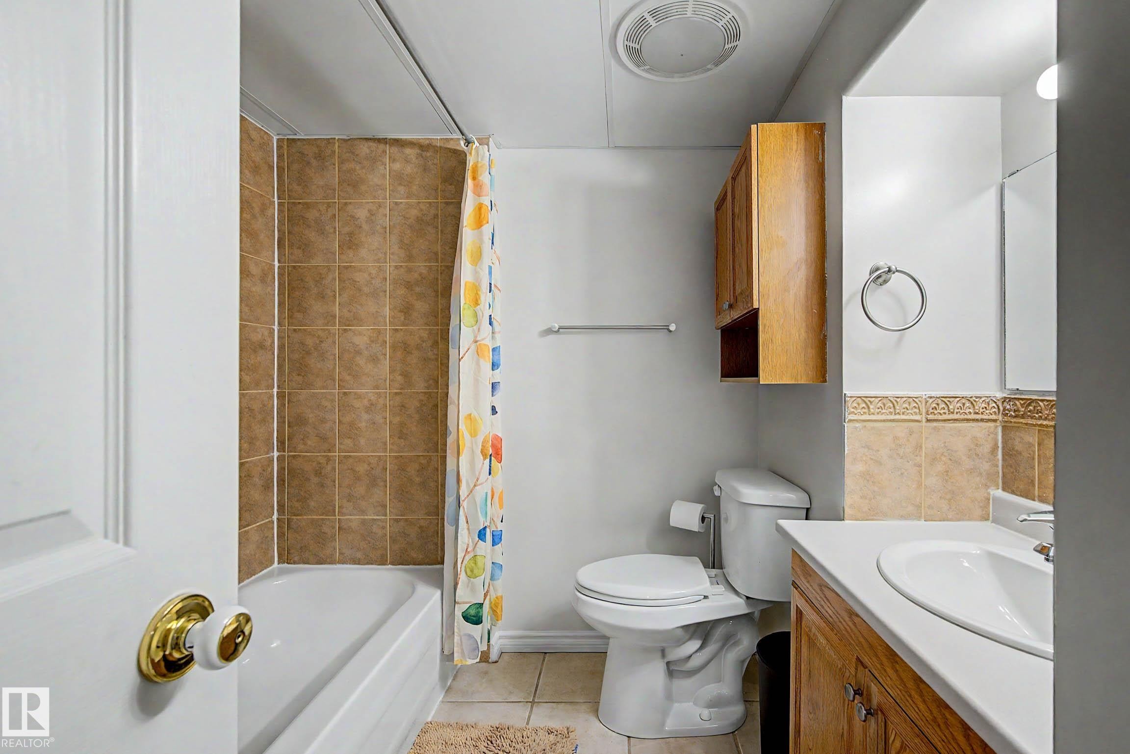 Bathroom featuring a tub/shower combination with brown square tile surround, a single vanity with an integrated sink and wood-finish cabinetry, a toilet, and a wall-mounted wood-finish cabinet - 3718 103B Street Nw, Edmonton, AB - Indoor Photo Showing Bathroom