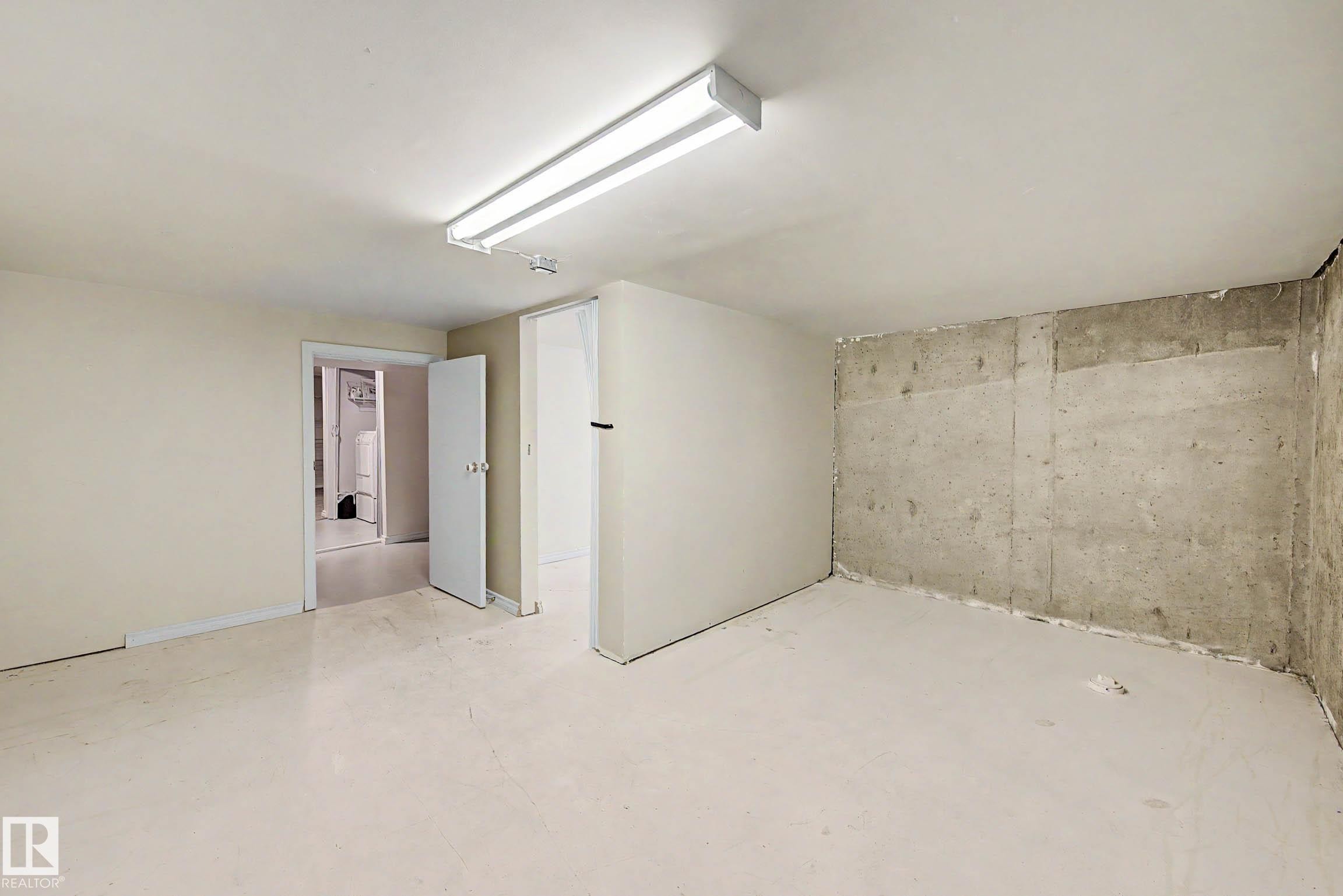 Spacious lower-level room featuring a concrete wall, neutral painted walls, light-toned flooring, and overhead fluorescent lighting - 3718 103B Street Nw, Edmonton, AB - Indoor