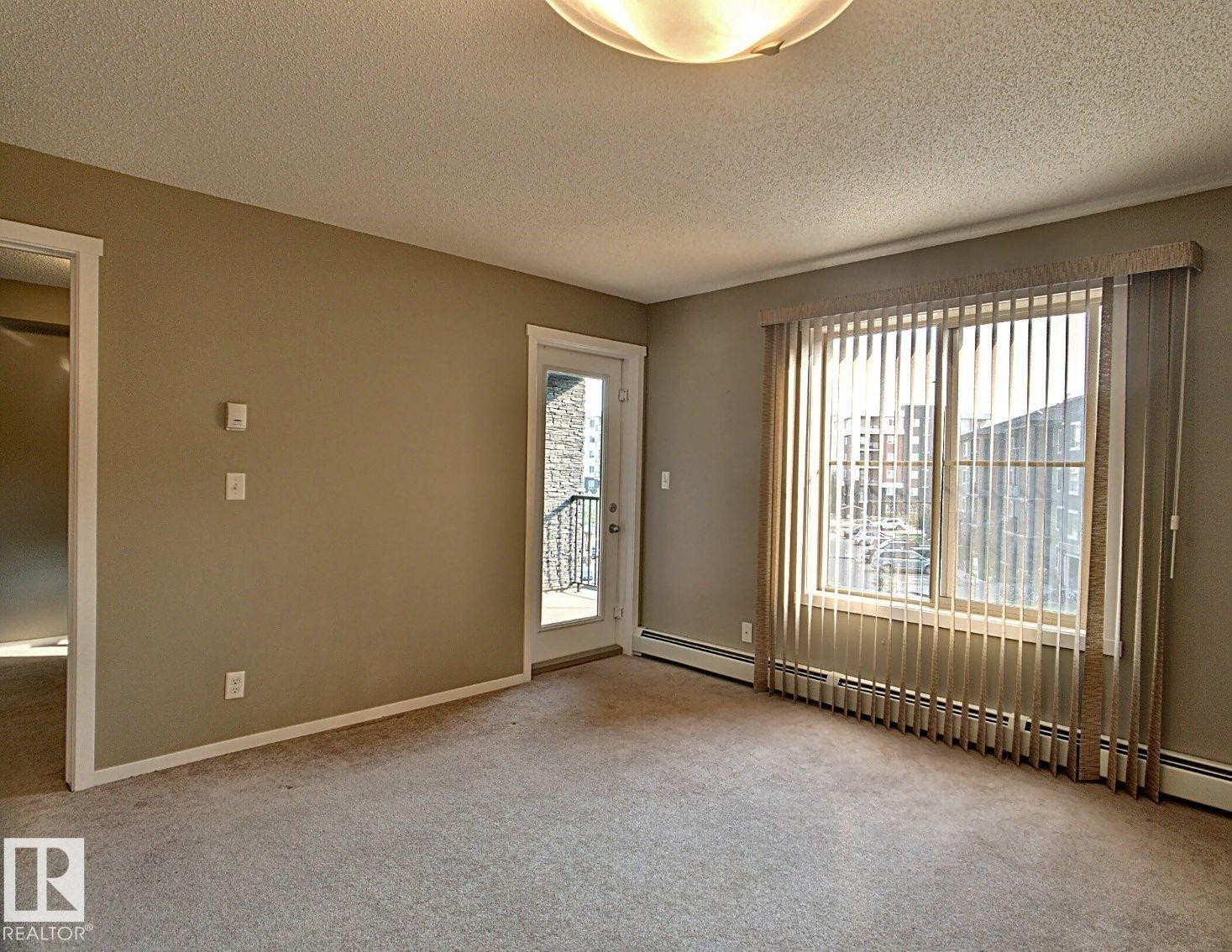 Neutral-toned carpeted room featuring a large window with vertical blinds, a glass-paneled door leading to a balcony with wrought iron railing, and a flush mount ceiling light fixture - 328 3315 James Mowatt Trail, Edmonton, AB