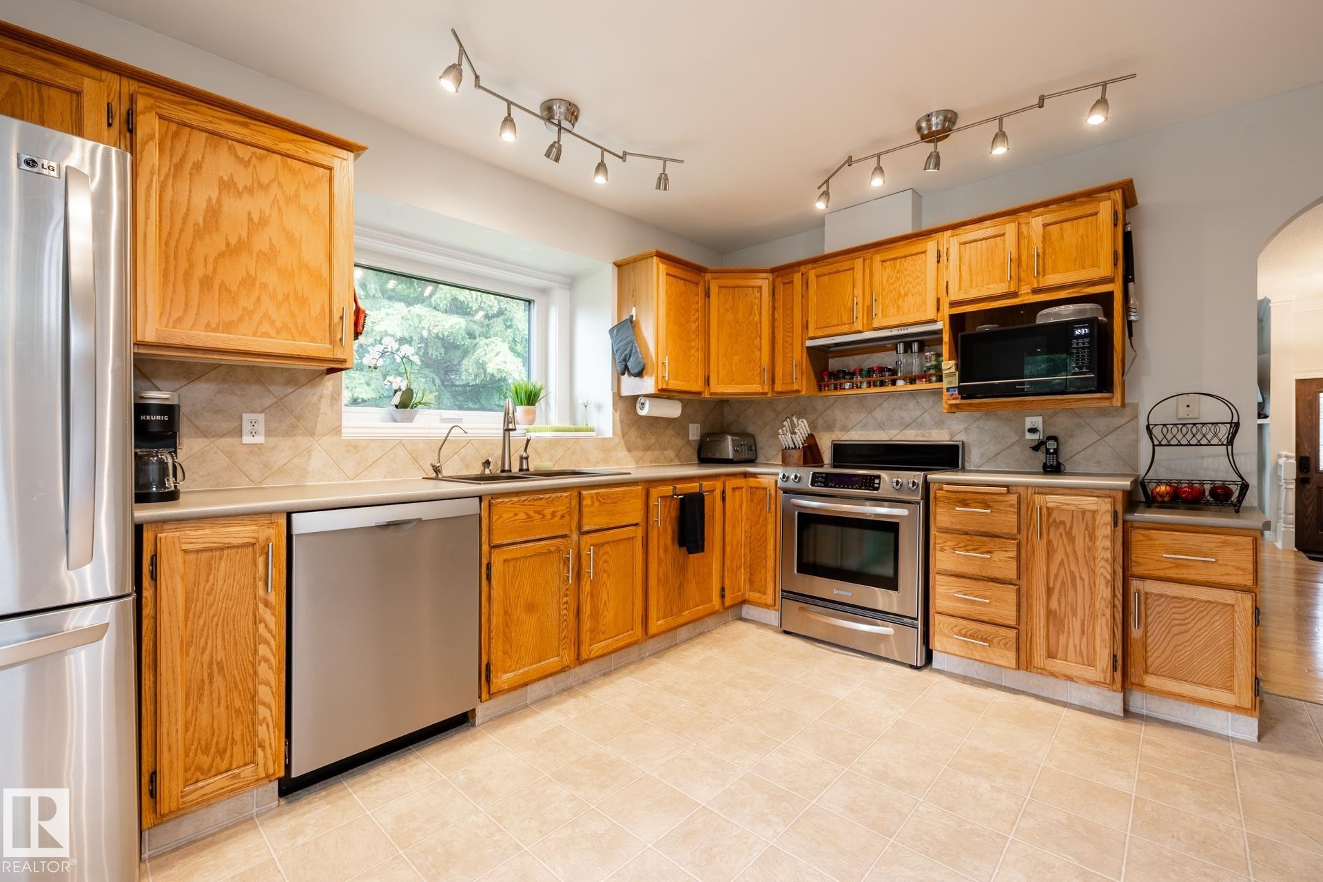 Kitchen featuring wood cabinetry, stainless steel appliances, track lighting, a tile backsplash, and a window above the sink - 27 Danforth Crescent, St. Albert, AB