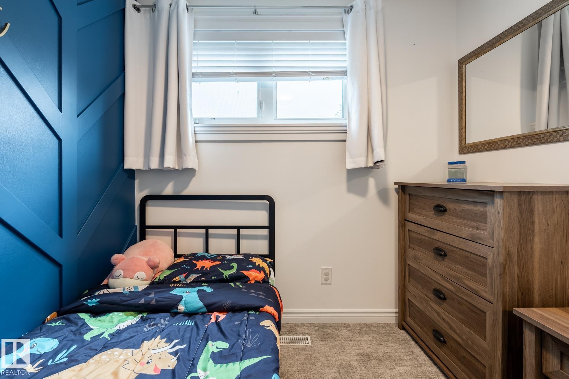 Accent wall with geometric paneling, wood-finish dresser with black hardware, window with white blinds, light gray wall, and gray carpeted flooring - 27 Danforth Crescent, St. Albert, AB