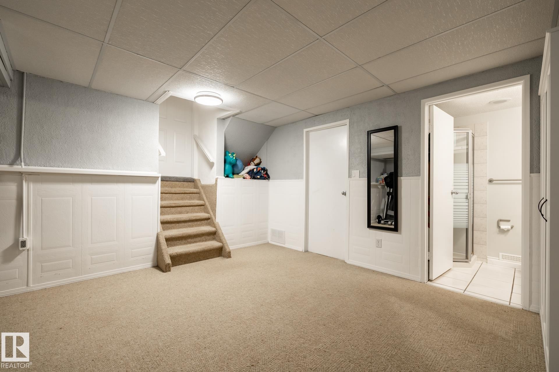 Carpeted room featuring light blue walls above white wainscoting, a drop ceiling, and a corner staircase - 27 Danforth Crescent, St. Albert, AB