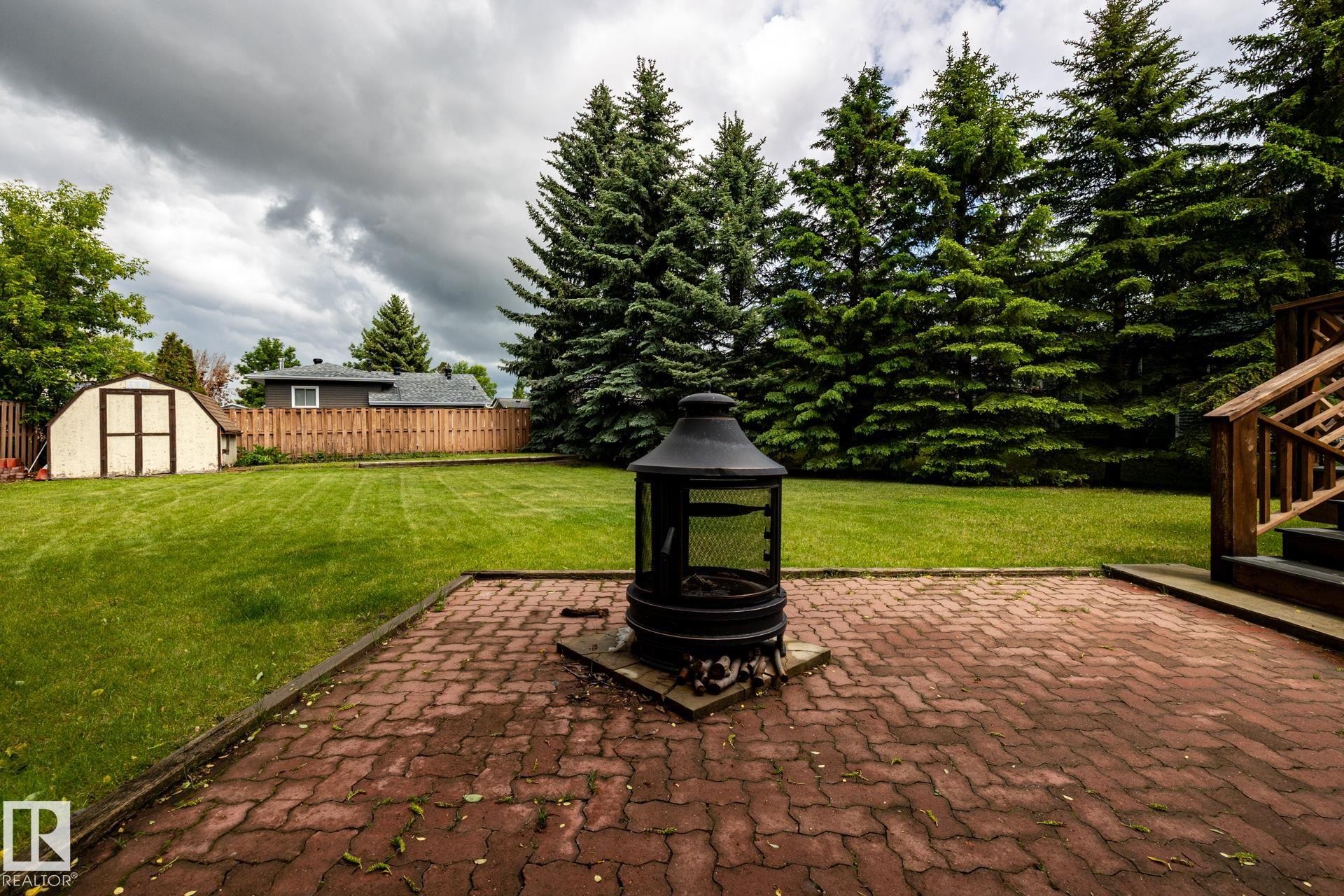 Expansive backyard featuring a red brick paver patio, a green lawn, mature evergreen trees, a wooden privacy fence, and a storage shed - 27 Danforth Crescent, St. Albert, AB