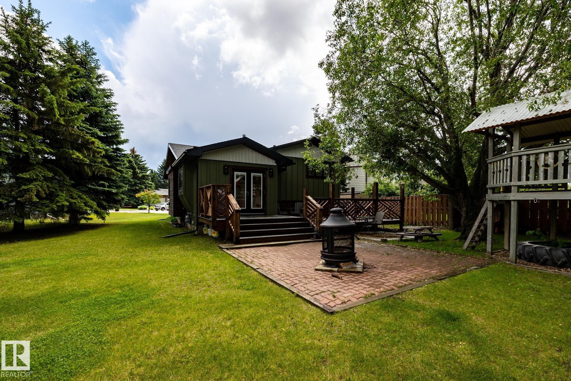 Expansive backyard featuring a red brick patio, green lawn, mature trees, and a wooden deck with railing - 27 Danforth Crescent, St. Albert, AB