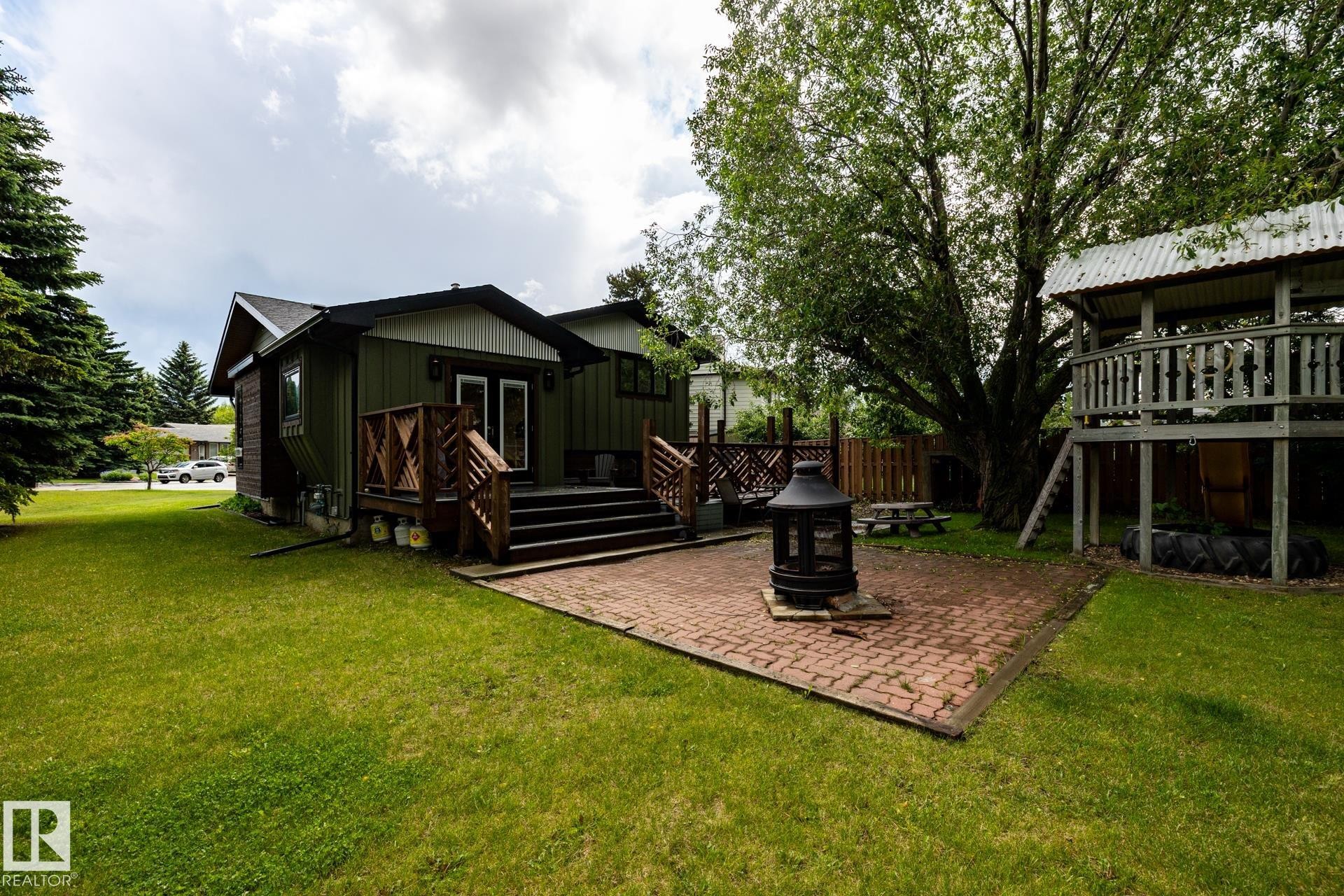 Spacious backyard featuring a brick paver patio with a central fire pit, extensive green lawn, and mature trees - 27 Danforth Crescent, St. Albert, AB