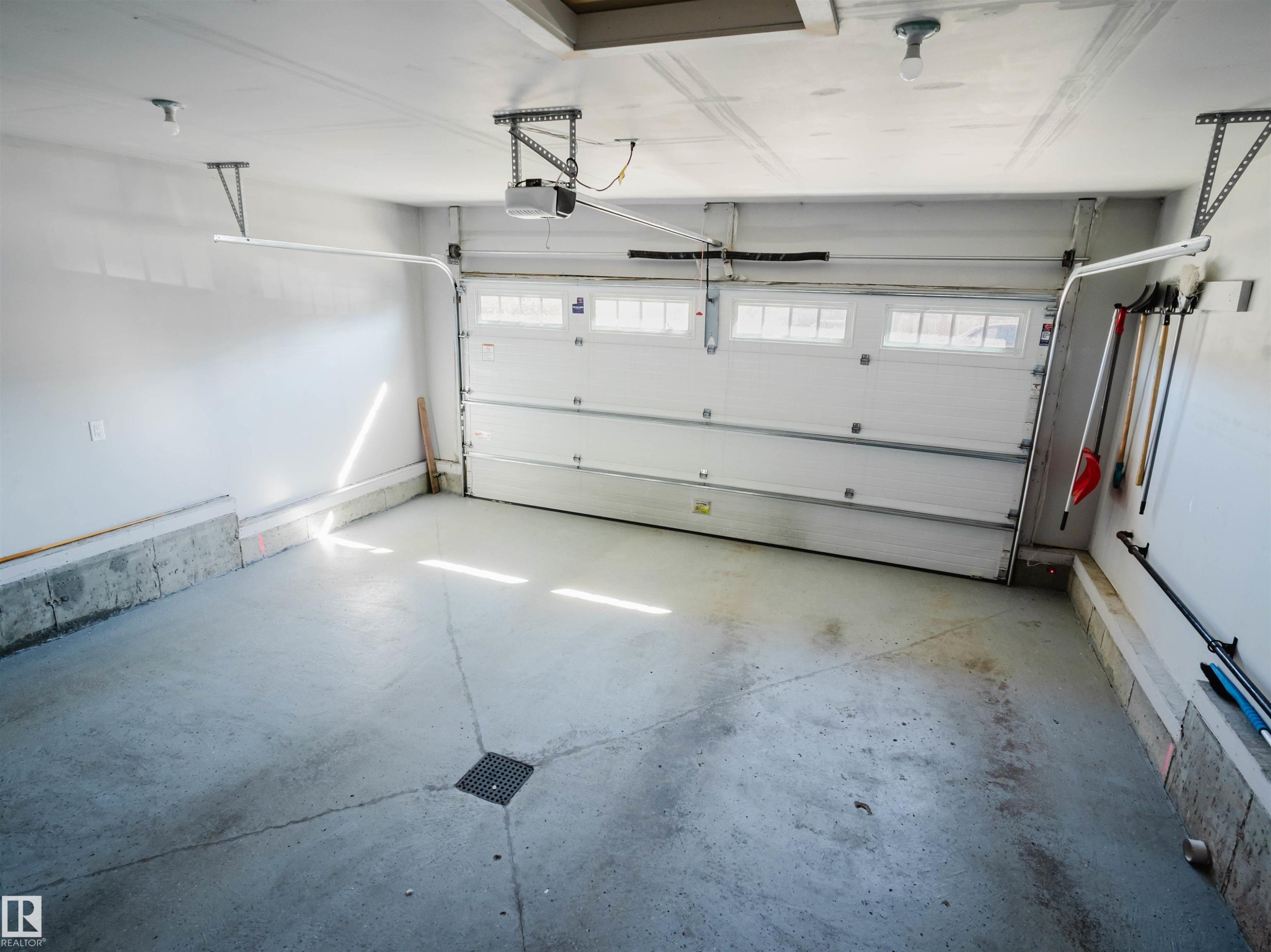 Spacious garage featuring a white overhead door with upper window panes, an automatic opener, a concrete floor with a central drain, and overhead lighting - 2526 Wonnacott Loop Sw, Edmonton, AB - Indoor Photo Showing Garage