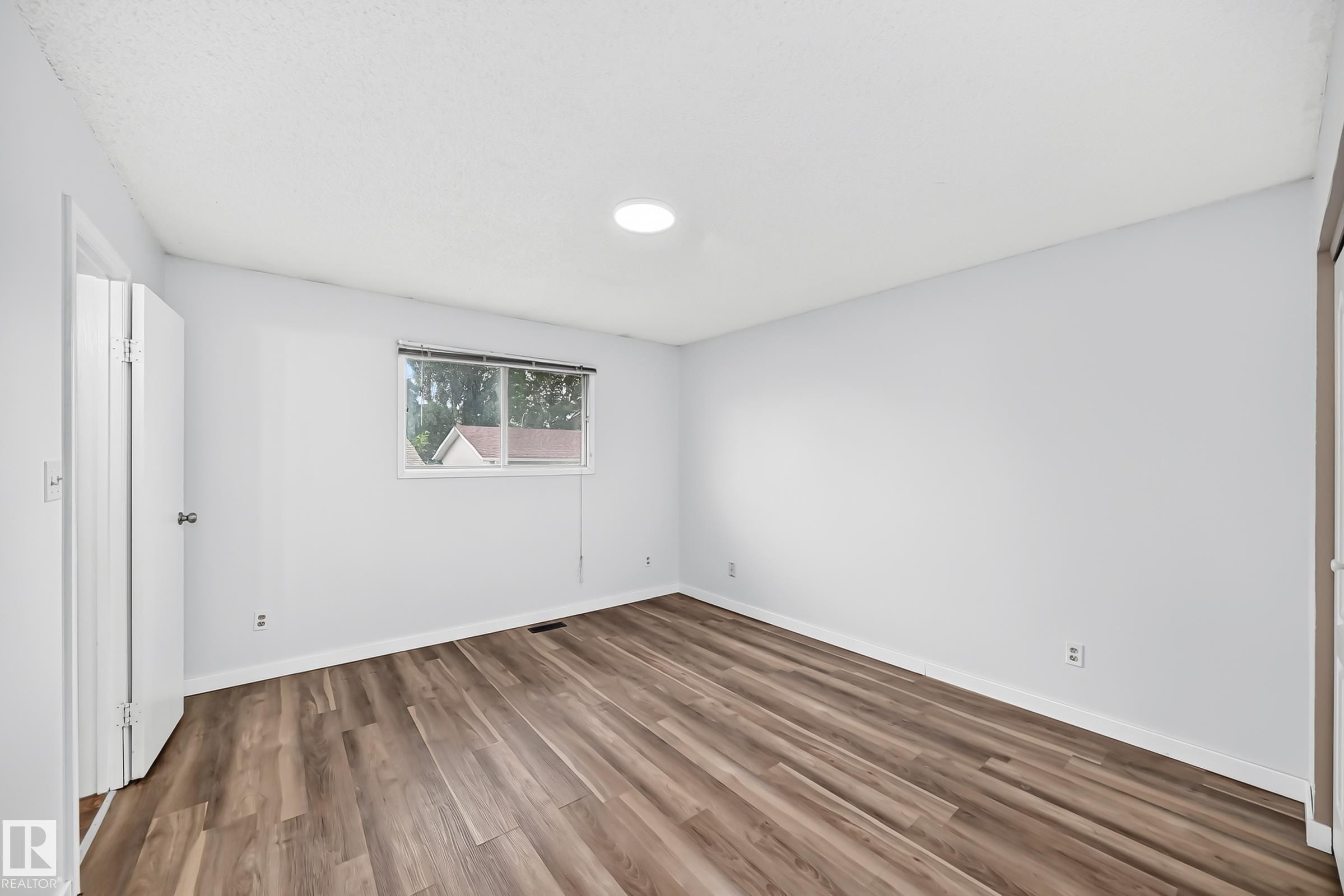 Spacious room featuring wide plank wood-finish flooring, a contemporary ceiling light fixture, a window with horizontal blinds, and white trim molding - 1103 56 Street, Edmonton, AB