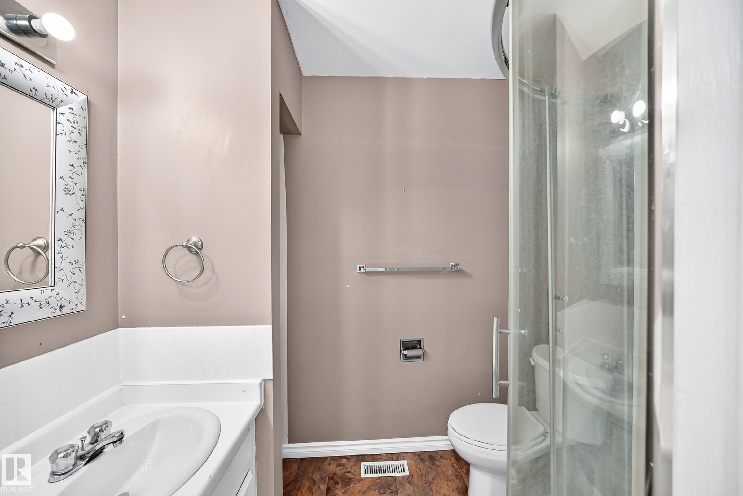 Bathroom featuring a white vanity with integrated sink, chrome faucet, mirrored medicine cabinet with decorative frame, glass-enclosed shower, and wood-finish flooring - 1103 56 Street, Edmonton, AB