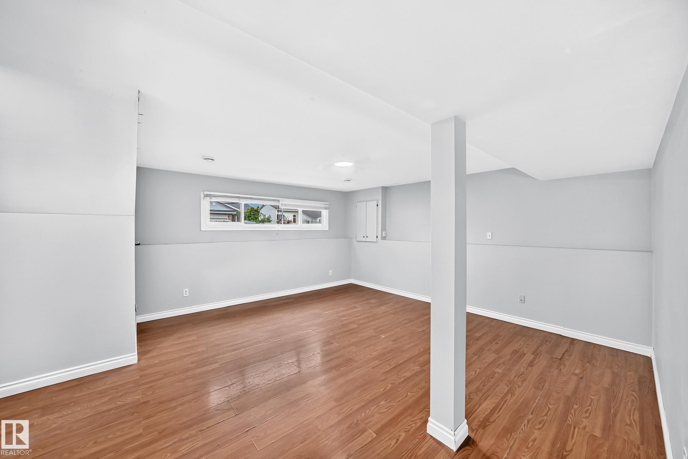 Spacious room featuring wood-finish flooring, light gray wall paint, white trim, and a window providing natural light - 1103 56 Street, Edmonton, AB