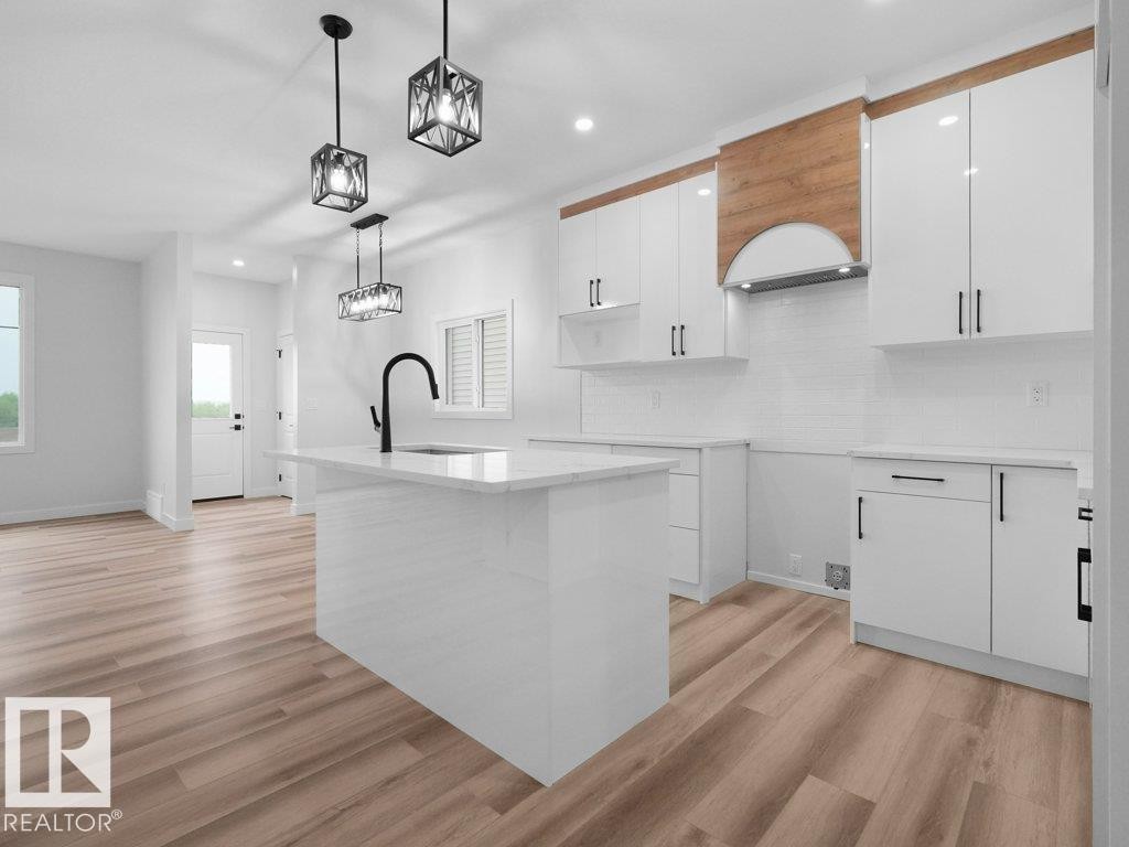 Modern kitchen featuring white cabinetry, a large center island with a black gooseneck faucet, wood-finish flooring, and a subway tile backsplash - 18719 130 Street Nw, Edmonton, AB