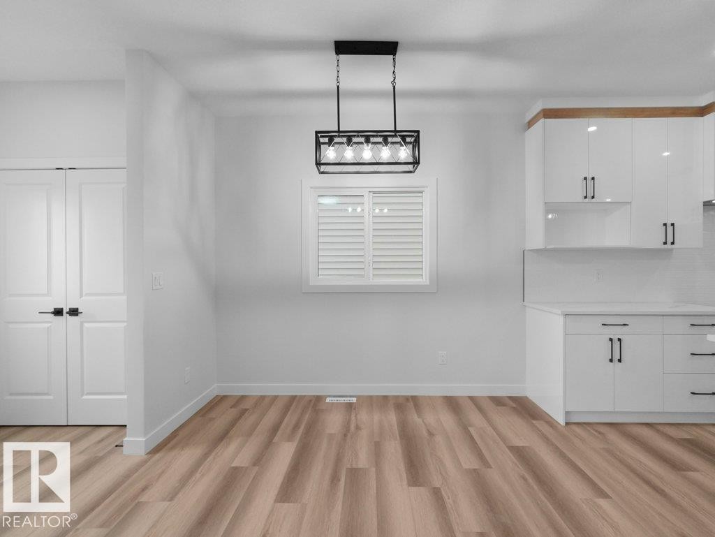 Open-concept room featuring wood-finish flooring, a contemporary linear chandelier, white plantation shutters, and a kitchen area with white cabinetry and a subway tile backsplash - 18719 130 Street Nw, Edmonton, AB