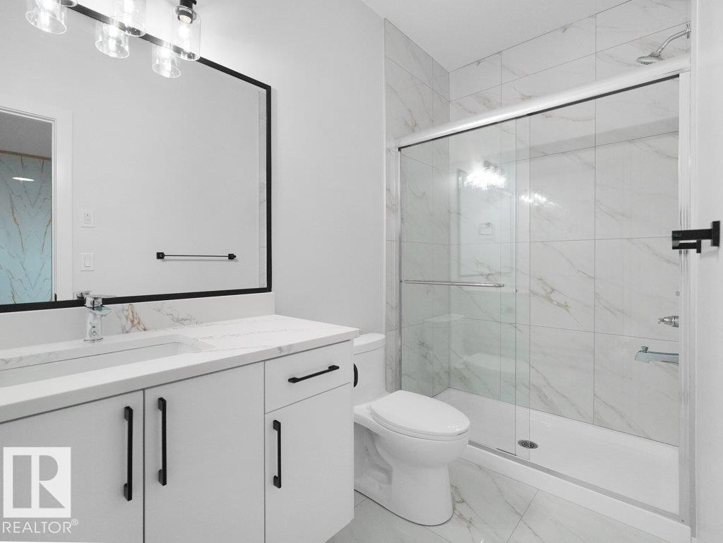 Bathroom featuring a glass-enclosed shower with white tiled walls and floor, a white vanity with a rectangular sink and stone countertop, a large framed mirror, and modern black hardware - 18719 130 Street Nw, Edmonton, AB