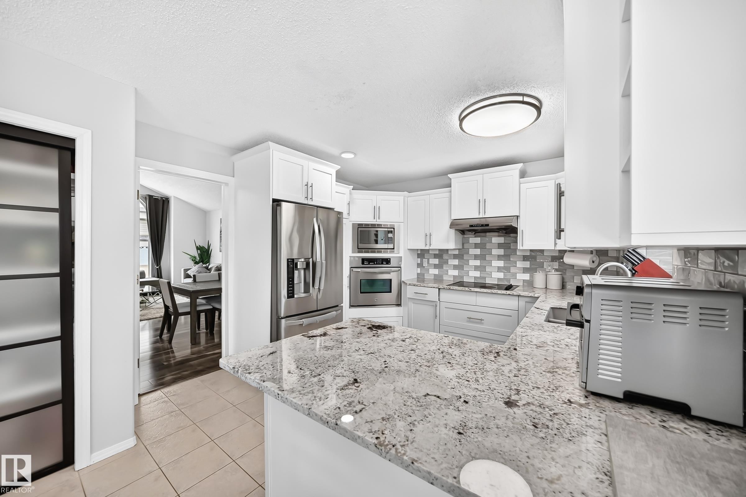Modern kitchen featuring white cabinetry, stainless steel appliances, a light-toned patterned backsplash, and a large breakfast bar with a stone-finish countertop - 4216 47 Street, Edmonton, AB