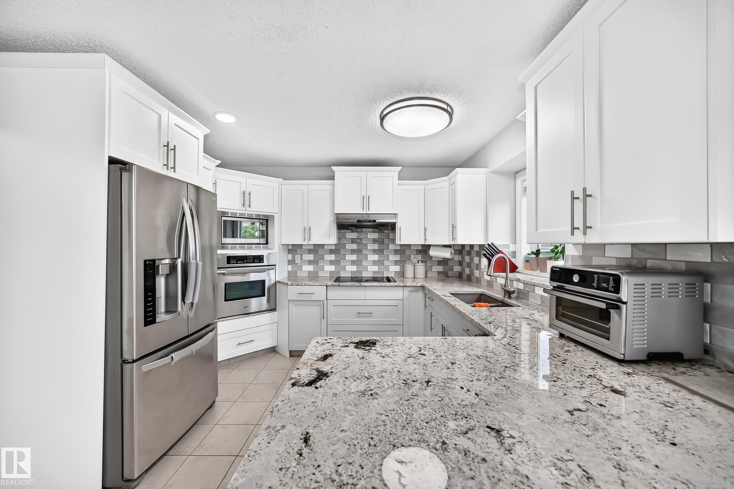 Kitchen featuring white shaker cabinetry, stainless steel appliances, a tile backsplash with a varied pattern, and speckled countertops - 4216 47 Street, Edmonton, AB