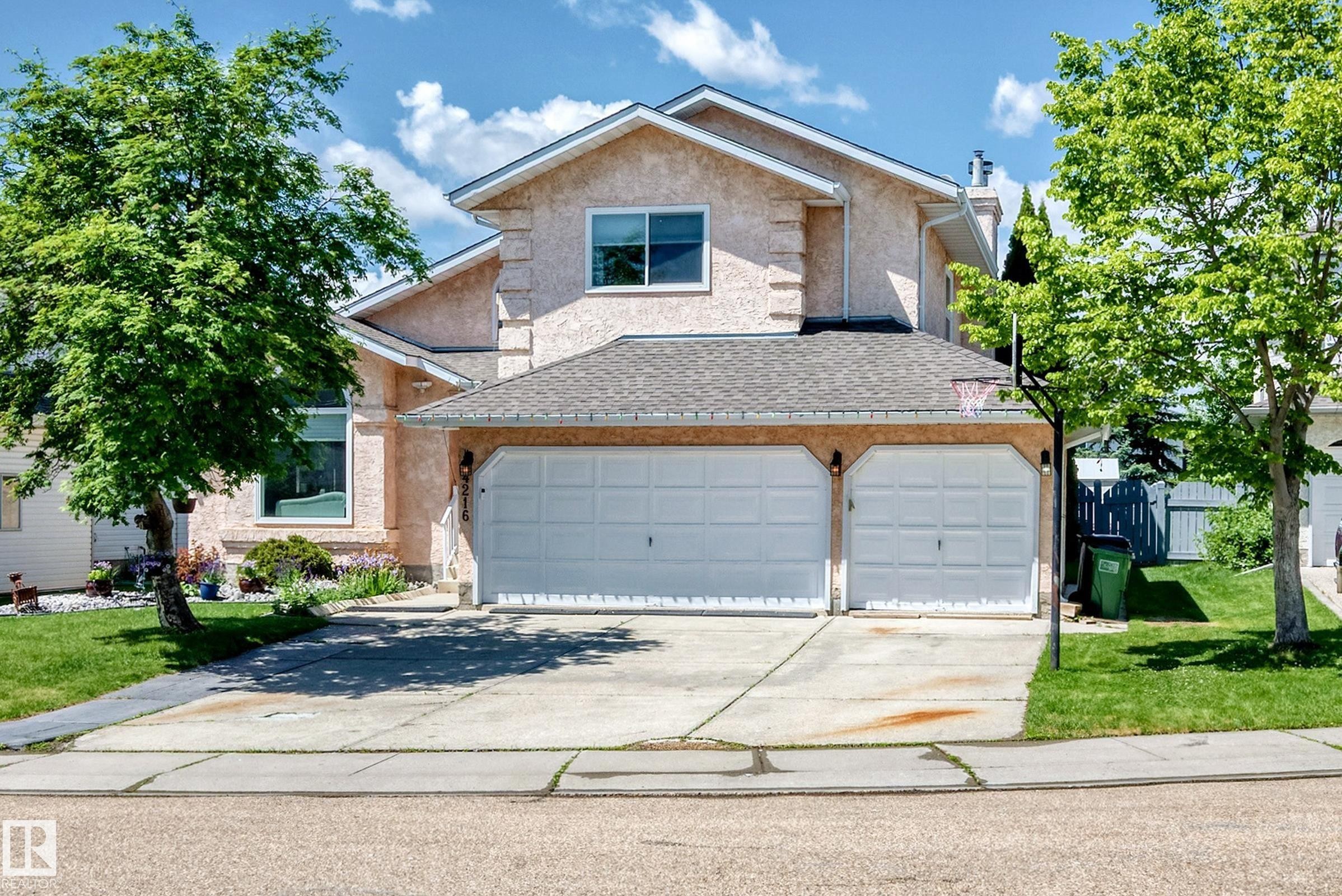 Stucco exterior with a peaked roofline, featuring two garage doors, a paved driveway, and mature landscaping - 4216 47 Street, Edmonton, AB