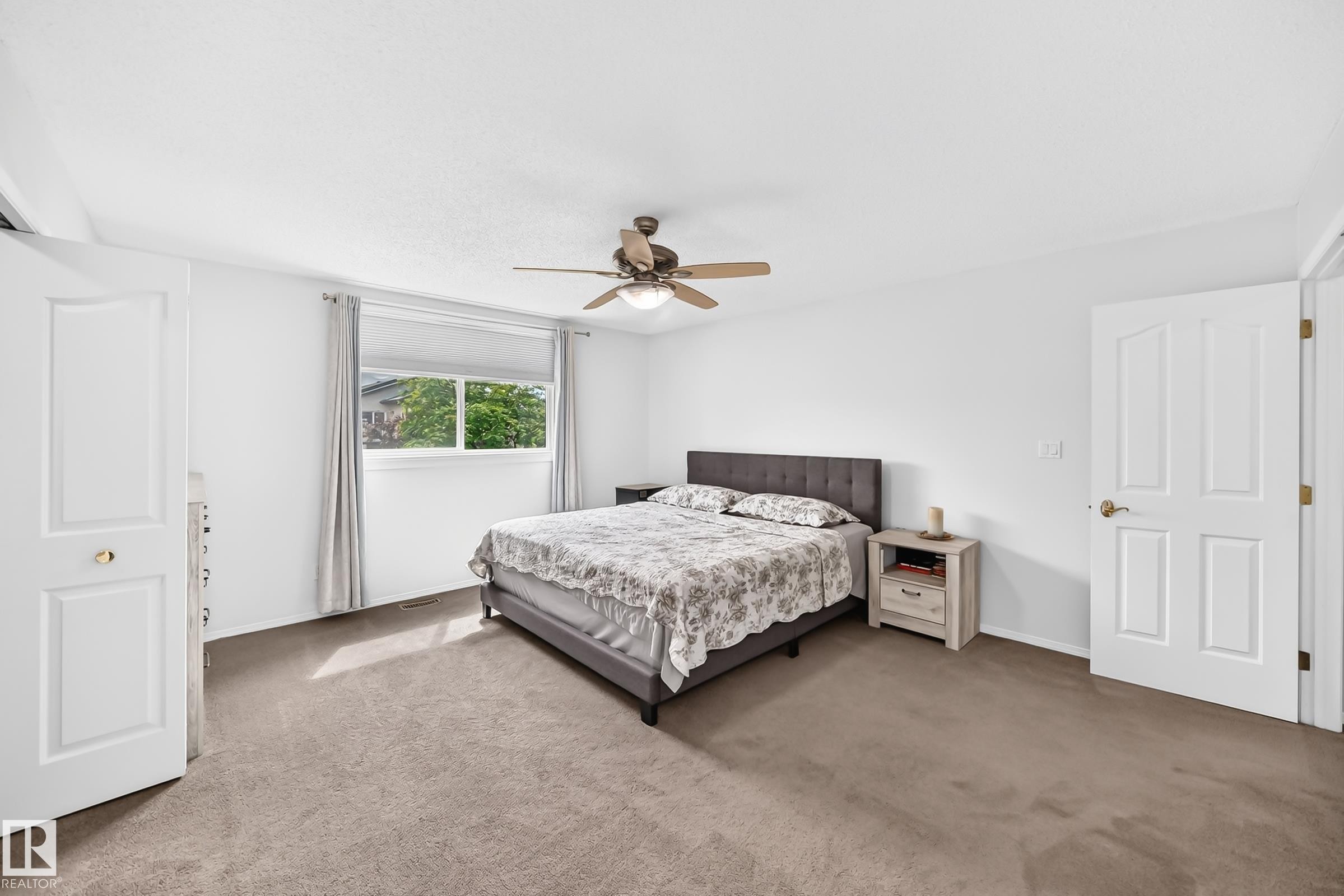 Spacious room featuring neutral carpeting, a ceiling fan with integrated lighting, and a large window with roller shades - 4216 47 Street, Edmonton, AB