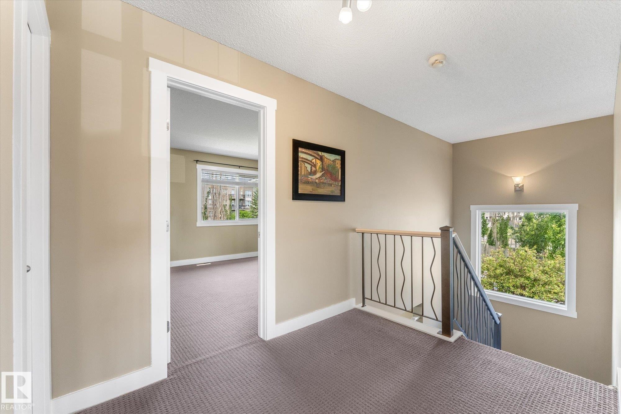 Carpeted upper-level landing featuring a wrought iron and wood handrail, a sconce wall light fixture, and a window overlooking exterior greenery - 488 Macewan Road, Edmonton, AB