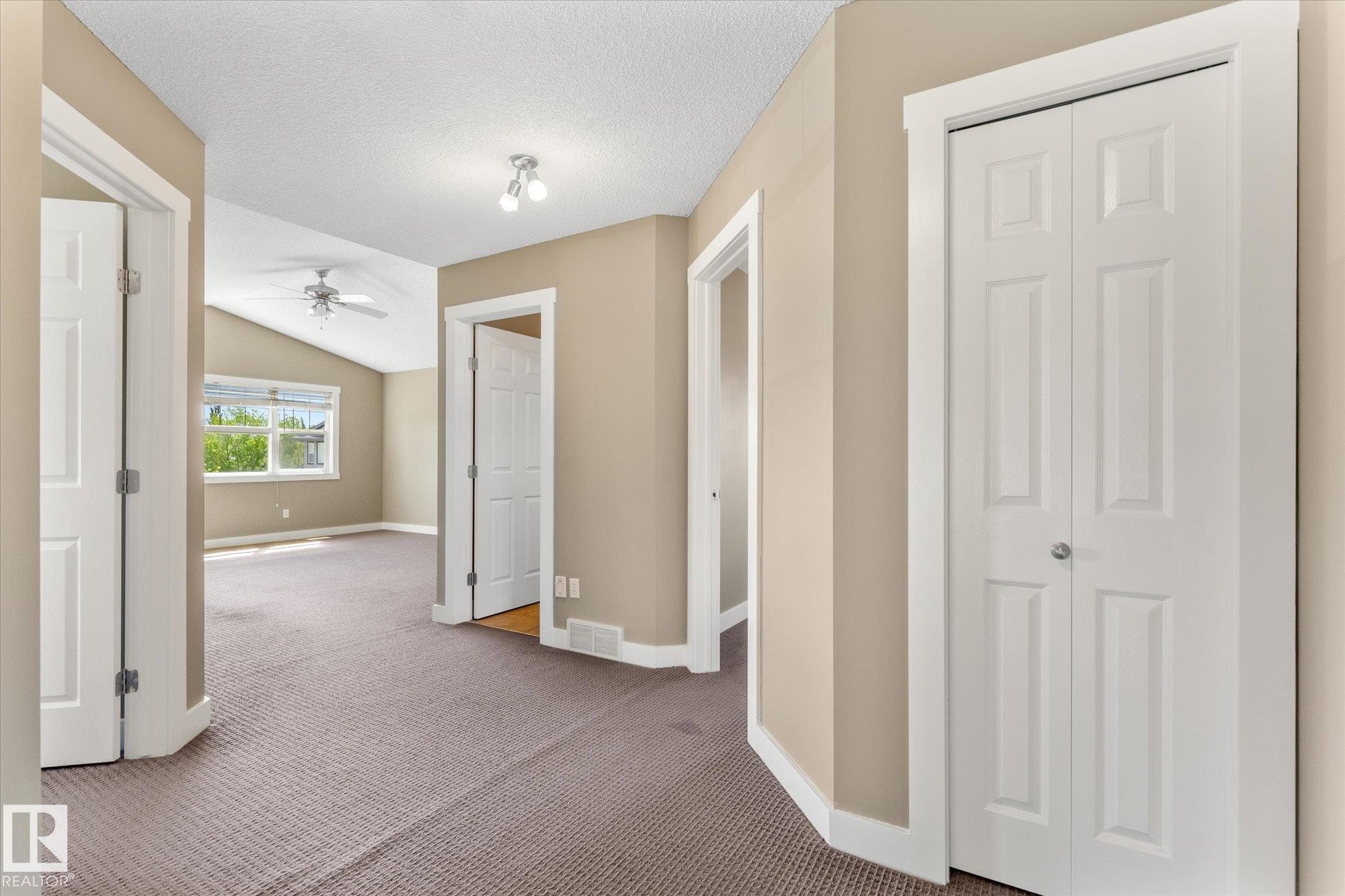 Carpeted hallway with three paneled doors and a bi-fold closet, leading to a room with vaulted ceilings, a ceiling fan, and large windows - 488 Macewan Road, Edmonton, AB