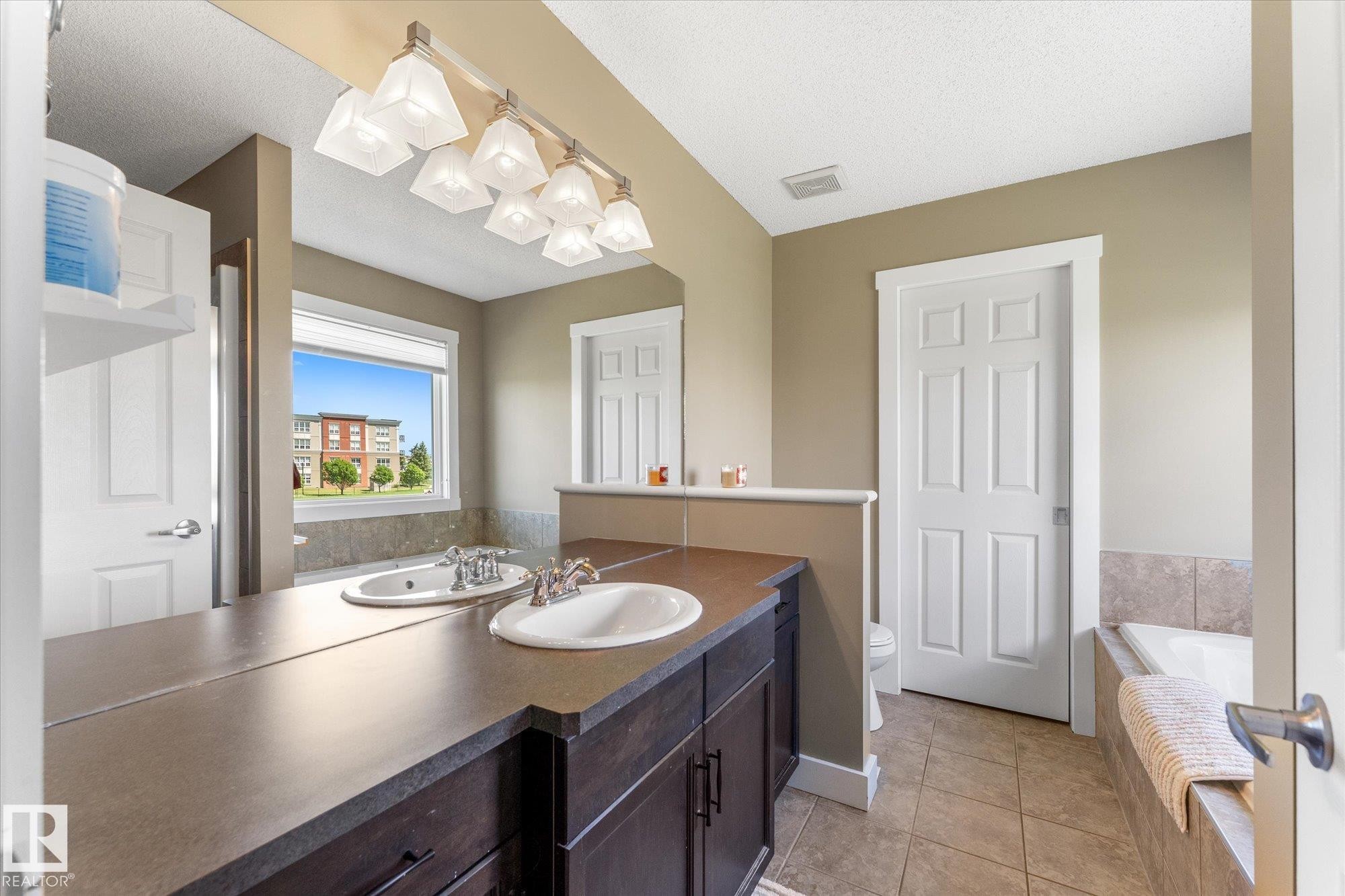 Bathroom featuring a double vanity with dark wood-finish cabinetry and integrated sinks, a wide window above a built-in tub with stone-finish tile surround, and neutral-toned wall paint - 488 Macewan Road, Edmonton, AB