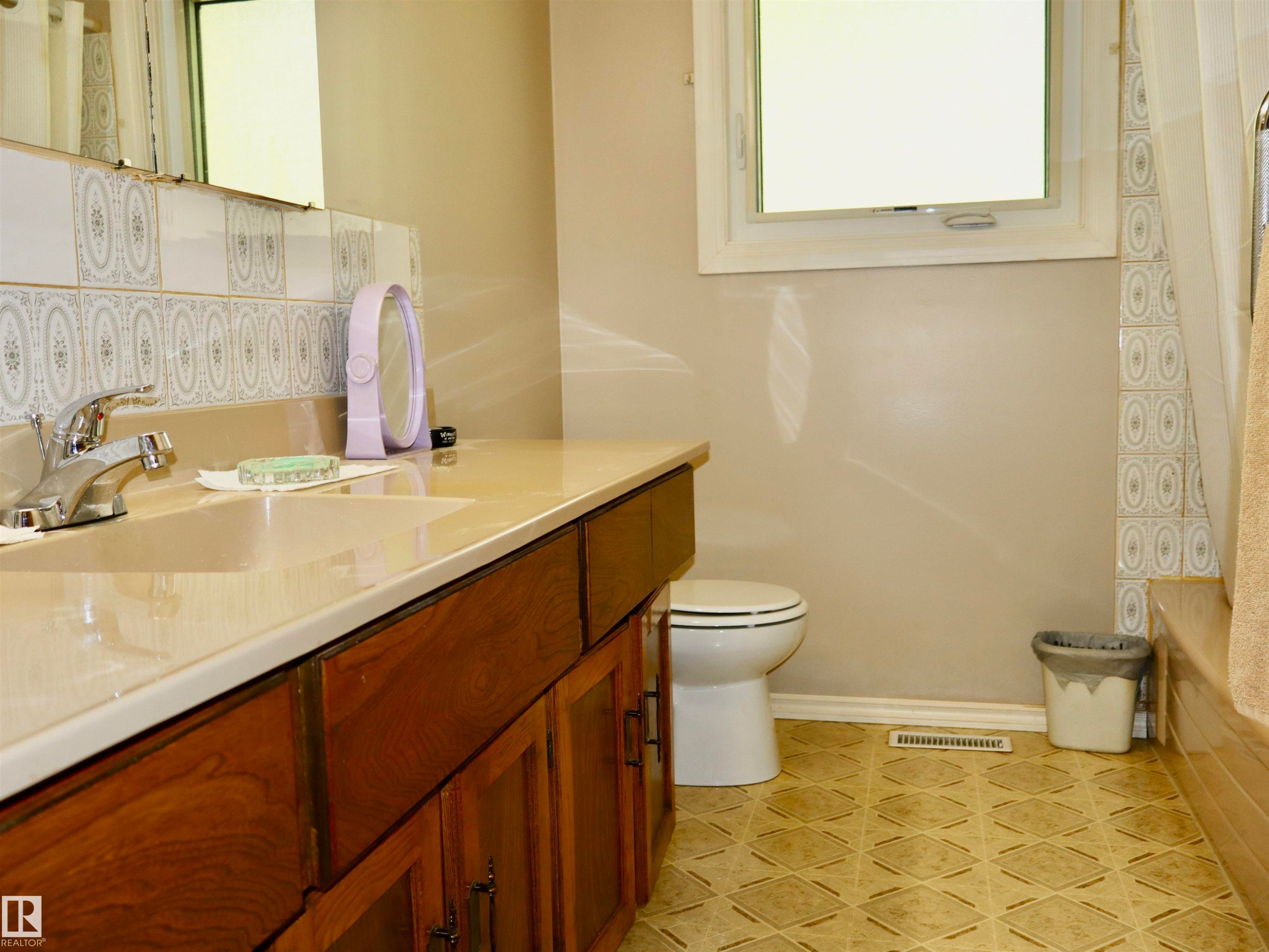 Bathroom featuring a wood-finish vanity with an integrated sink and countertop, patterned tile backsplash, and a window with white trim - 5530 46 Street, Barrhead, AB