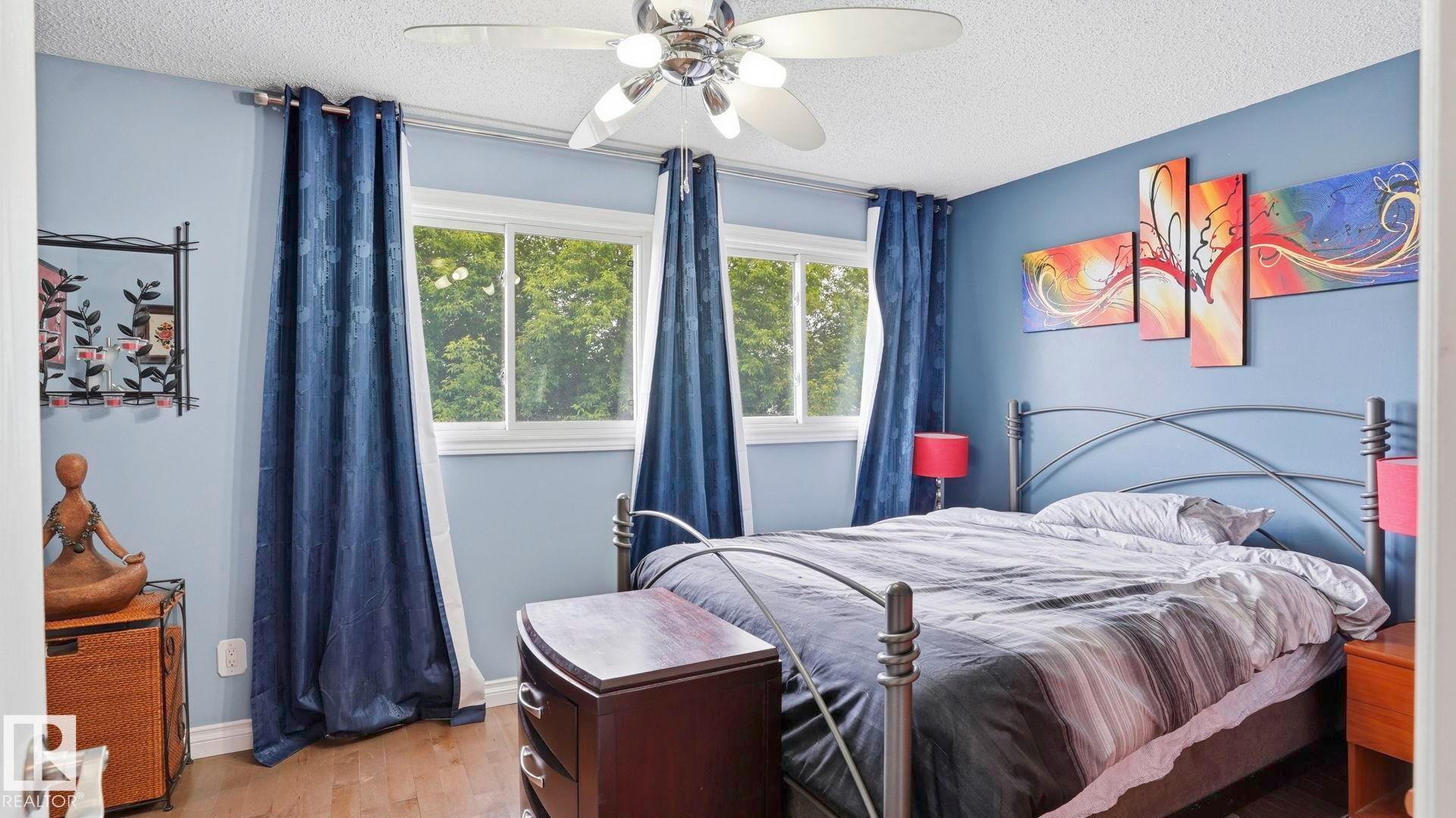 Bedroom featuring two windows with blue drapery, a ceiling fan with integrated lighting, wood-finish flooring, and blue walls - 1827 36 Street Nw, Edmonton, AB