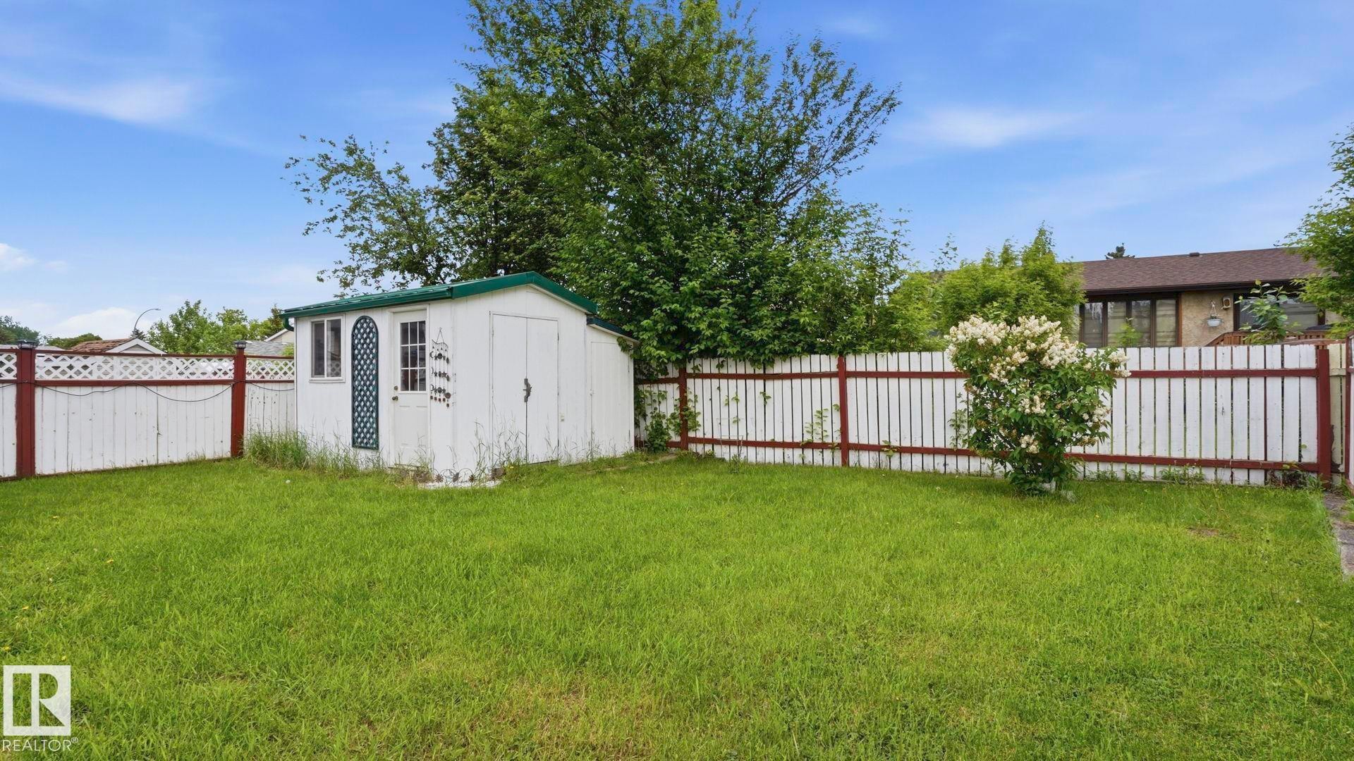 Grassed yard featuring a white wooden fence with a red top rail, mature trees, and a storage shed with a green roof - 1827 36 Street Nw, Edmonton, AB