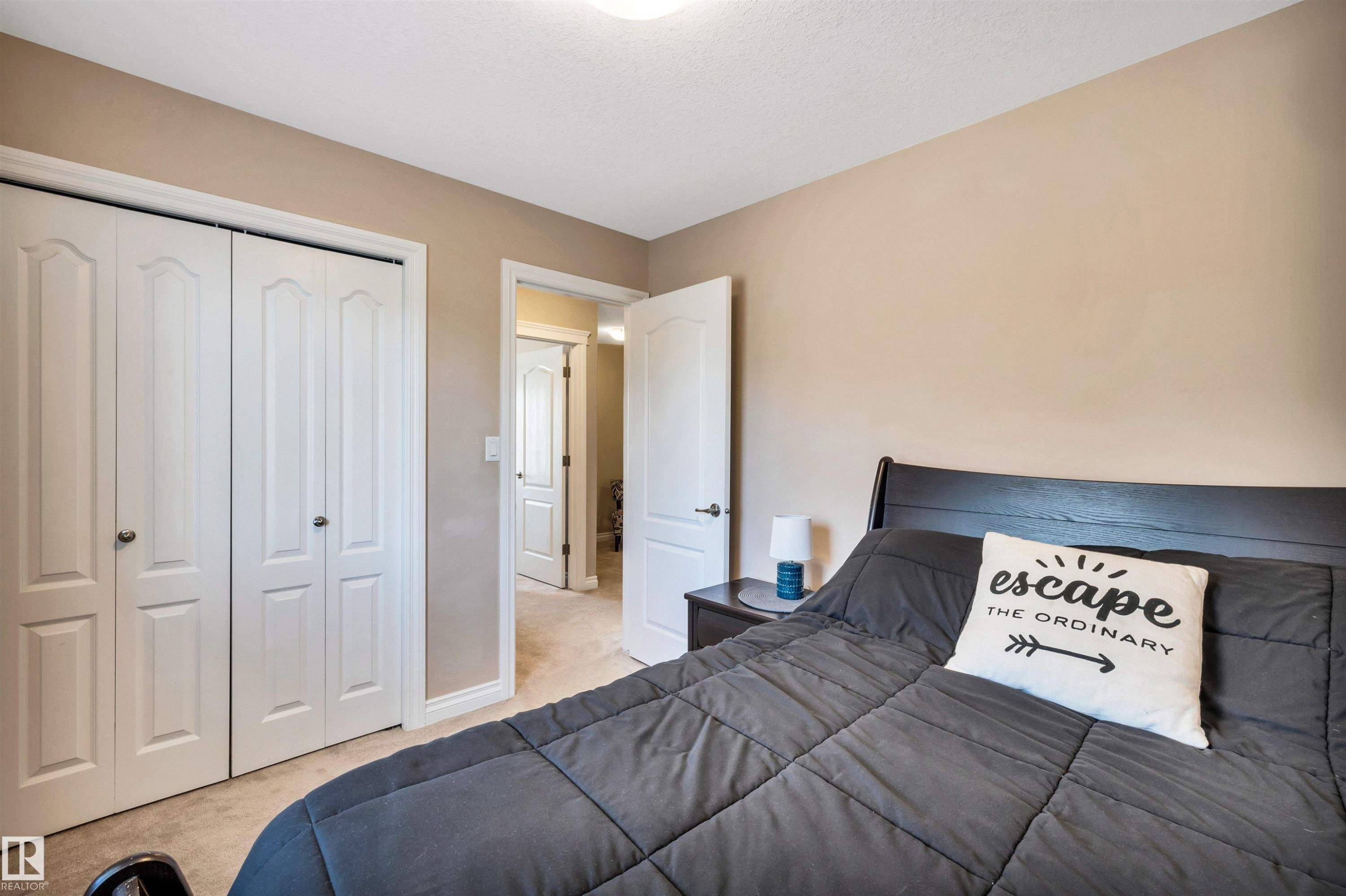 Neutral-toned bedroom featuring bi-fold closet doors, light-colored carpeting, and a contrasting dark wood-finish headboard - 18 Voleta Crest, Spruce Grove, AB