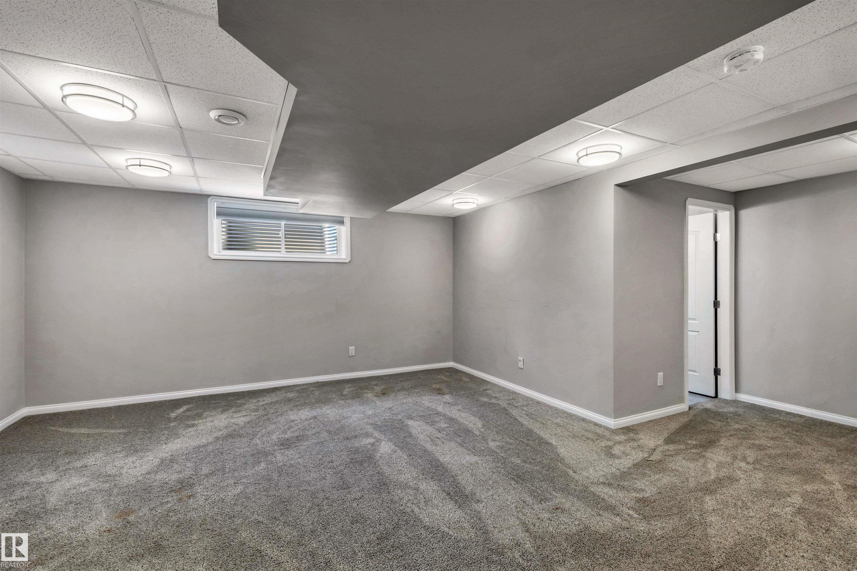Spacious lower-level room featuring plush gray carpeting, neutral wall finishes, and a combination of drop-panel and flat ceilings with flush-mount lighting fixtures - 18 Voleta Crest, Spruce Grove, AB