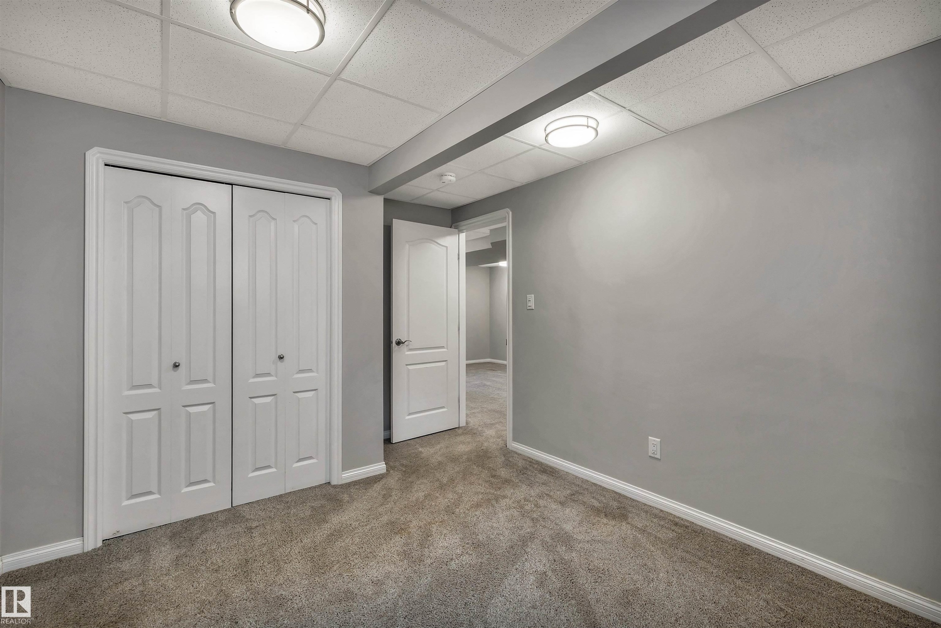 Room featuring light gray walls, gray carpet flooring, white bi-fold closet doors, white baseboards, and a drop ceiling with integrated lighting - 18 Voleta Crest, Spruce Grove, AB
