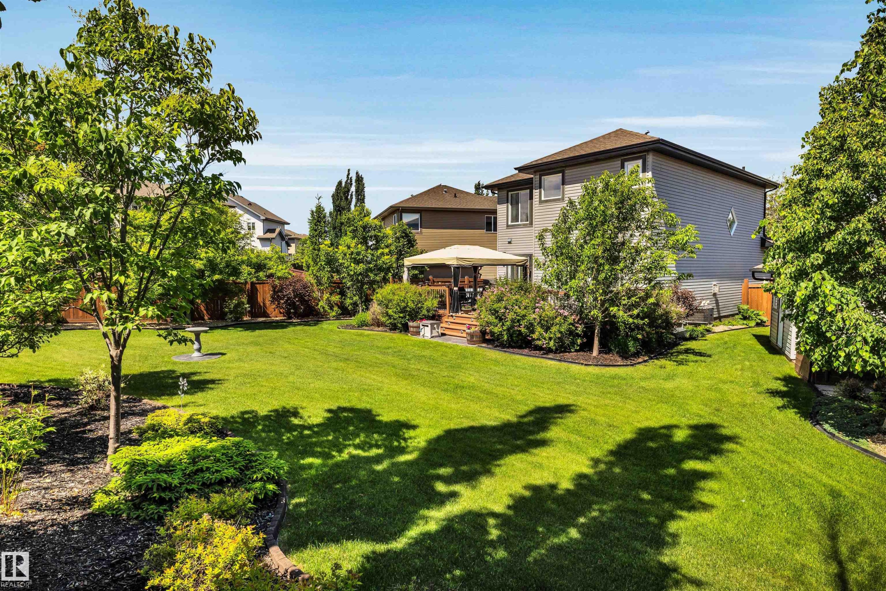Spacious backyard featuring a large green lawn, mature trees, landscaped garden beds, a wooden deck, and a privacy fence - 18 Voleta Crest, Spruce Grove, AB