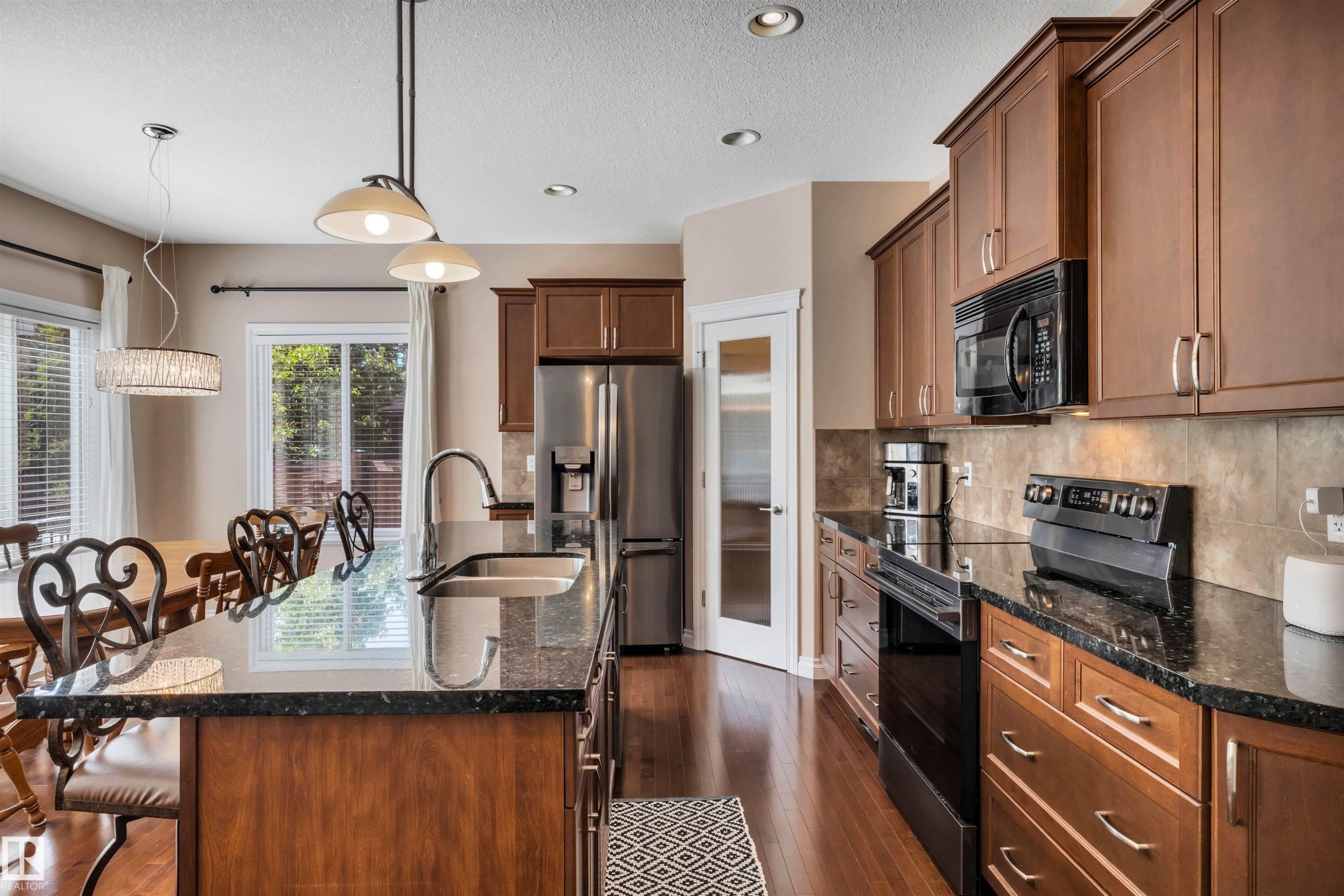 Contemporary kitchen featuring an expansive island with a double basin sink, dark stone countertops, wood-finish cabinetry, stainless steel appliances, and recessed lighting - 18 Voleta Crest, Spruce Grove, AB