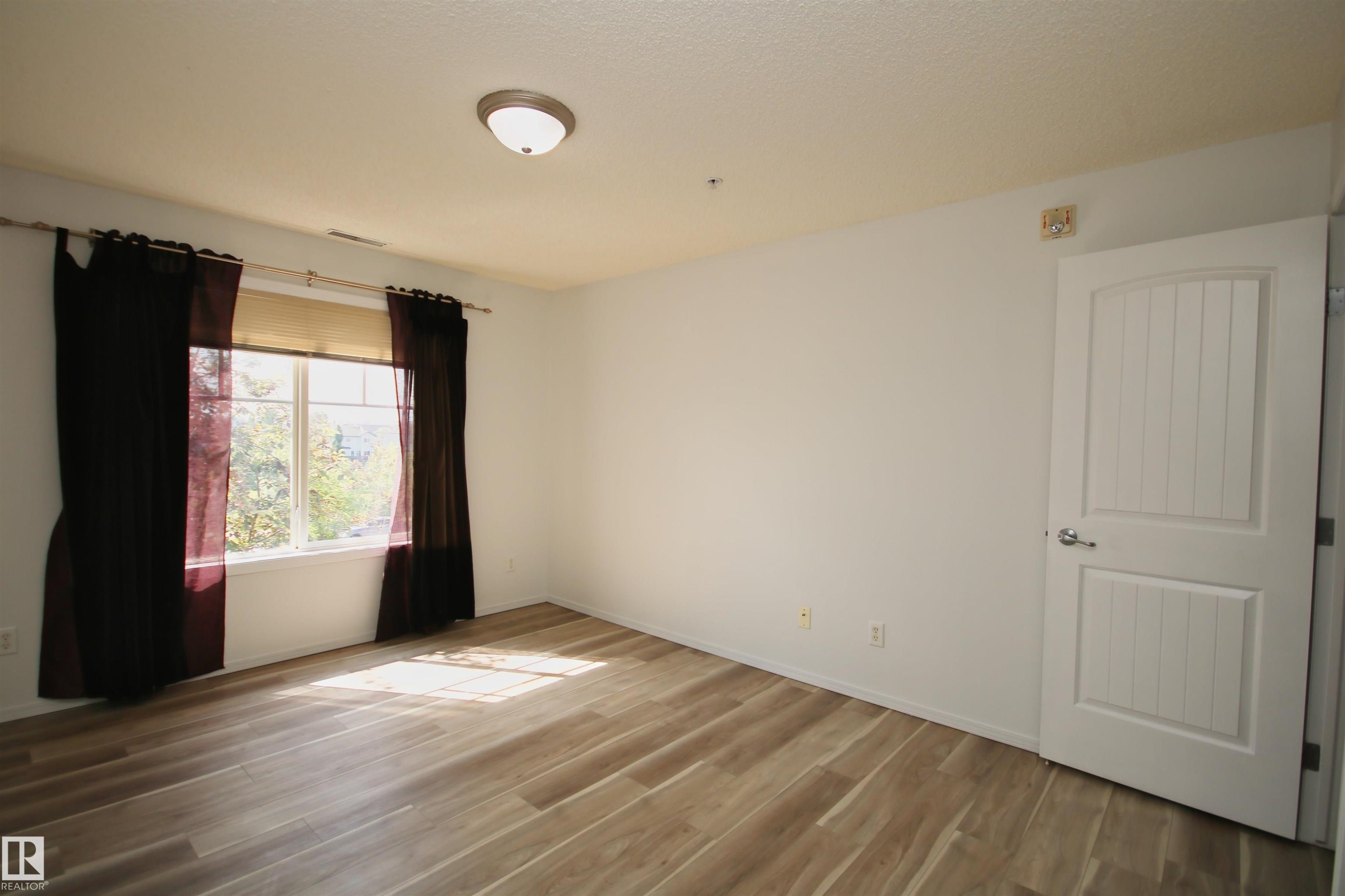 Spacious room featuring wood-finish flooring, a large window, and a white paneled door with brushed nickel hardware - 311 2098 Blackmud Creek Drive, Edmonton, AB