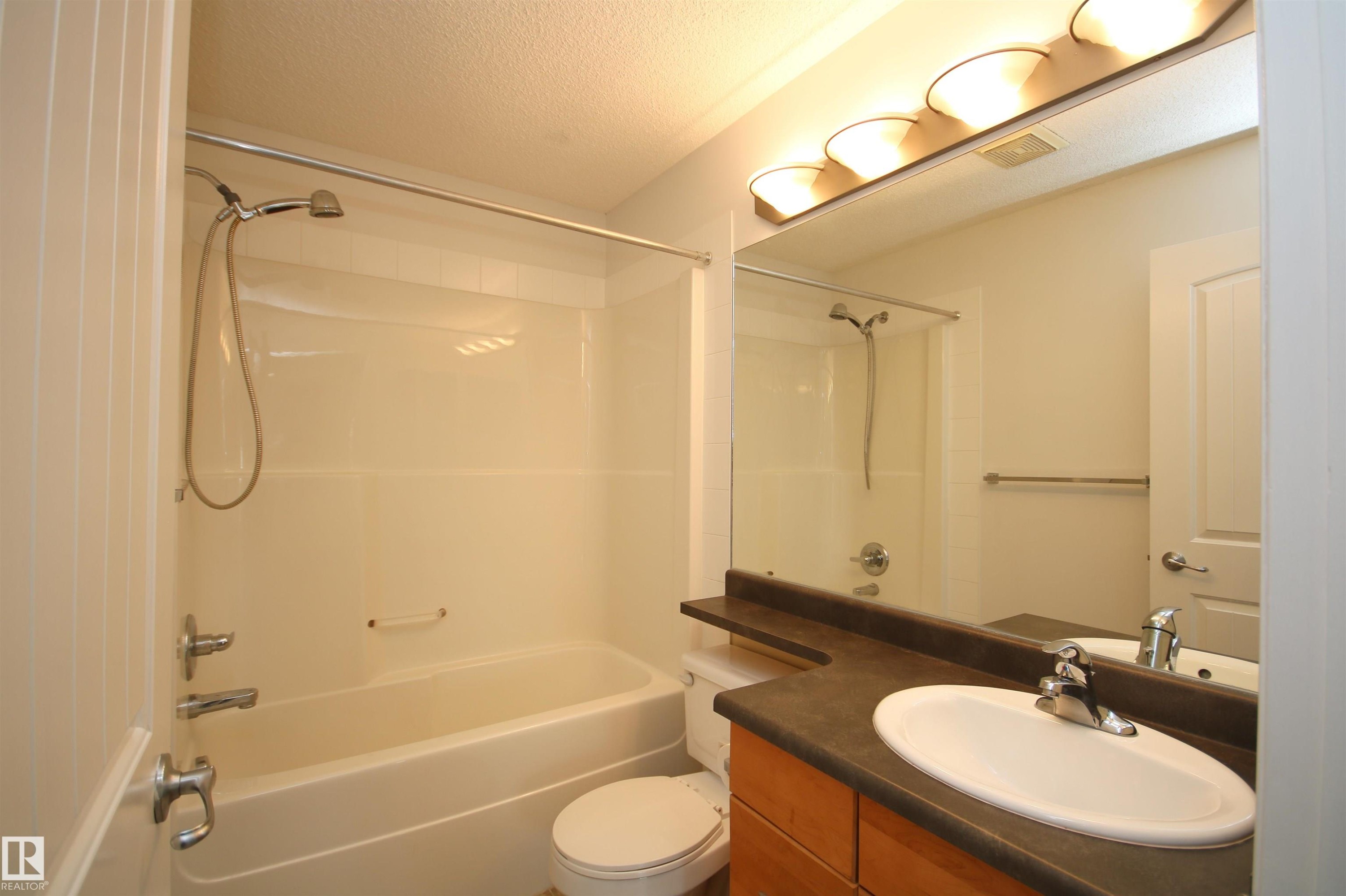 Bathroom featuring a combination bathtub/shower with a curved shower rod, a vanity with a dark countertop and integrated sink, and an overhead light fixture with multiple bulbs - 311 2098 Blackmud Creek Drive, Edmonton, AB