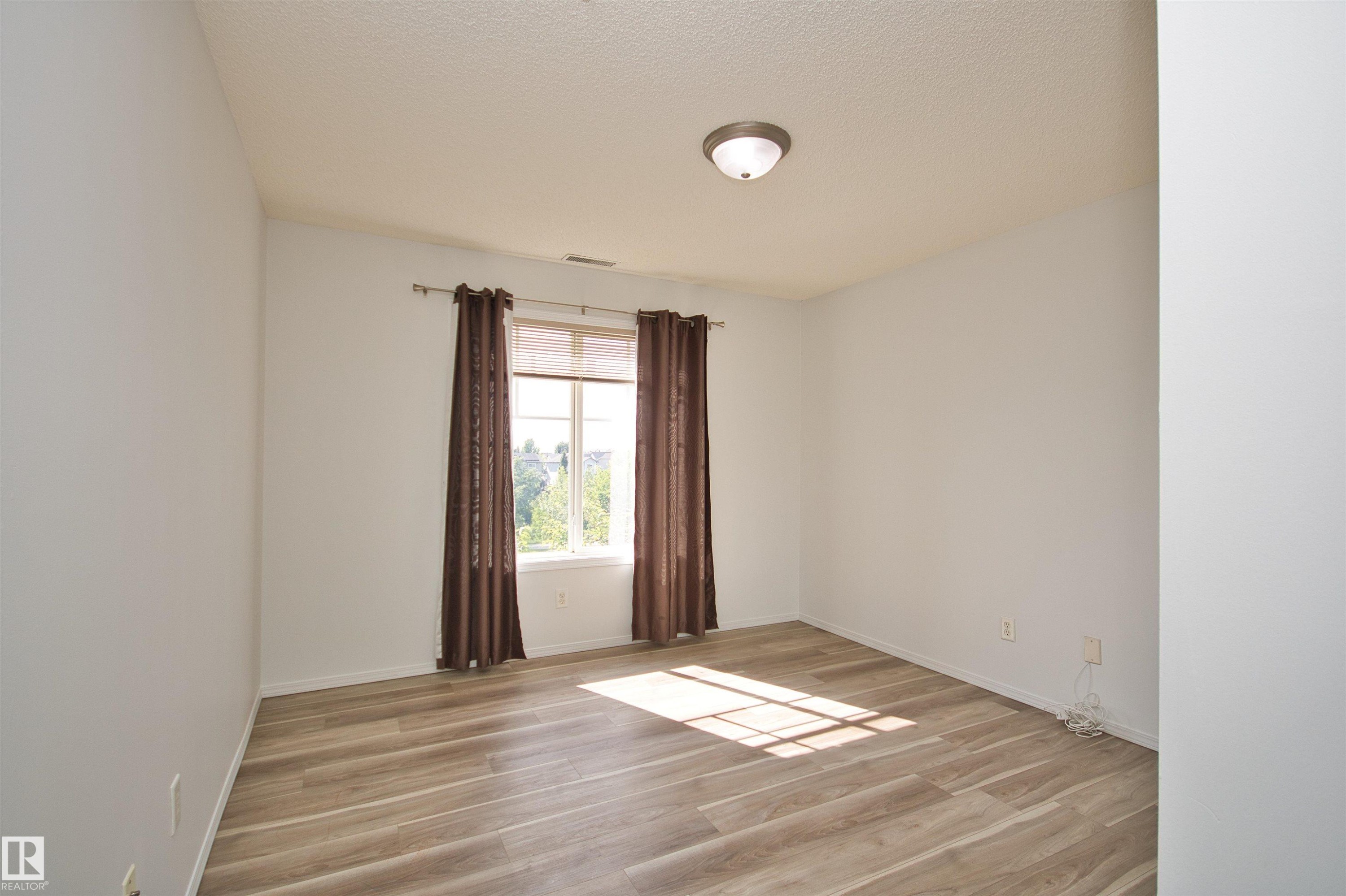 Spacious room with light wood-finish flooring, a contemporary flush-mount ceiling light fixture, and a large window providing ambient light - 311 2098 Blackmud Creek Drive, Edmonton, AB